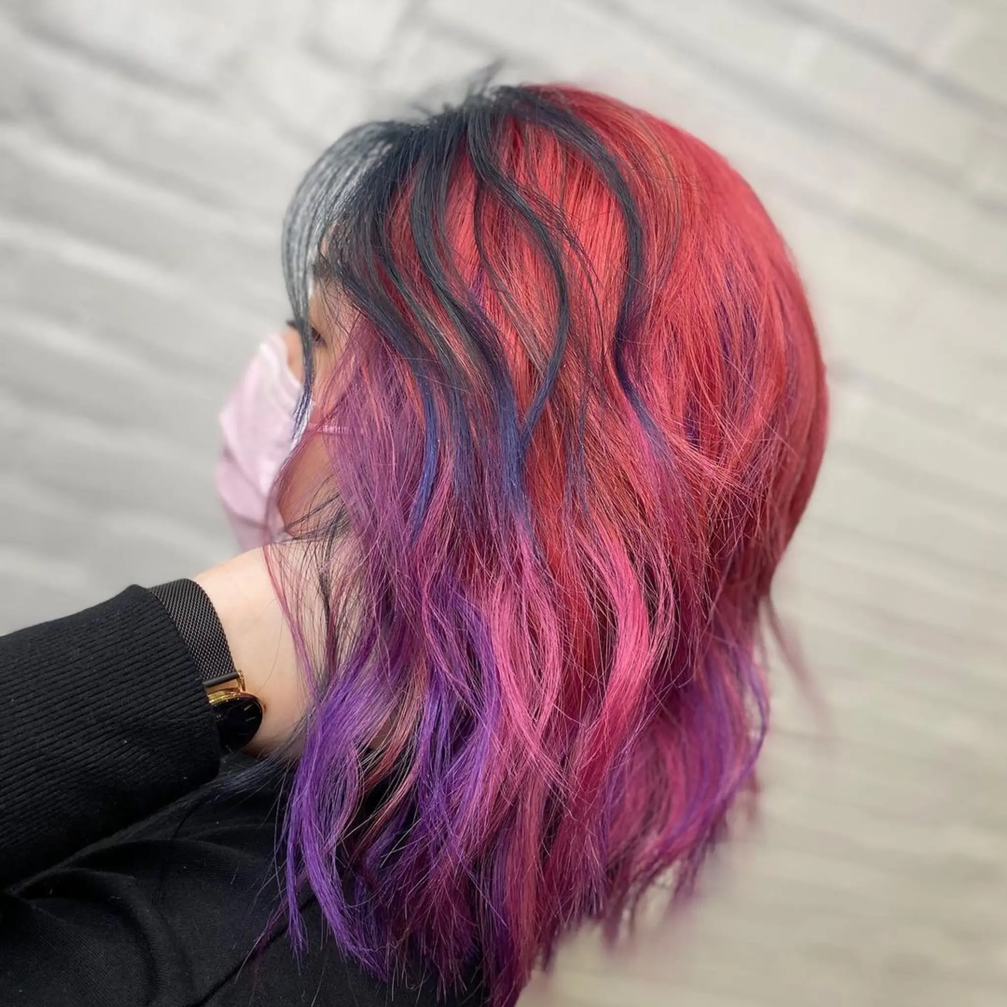 A medium-length wavy hairstyle with an ombre effect, featuring black roots and transitioning to vibrant red, pink, and purple hues.