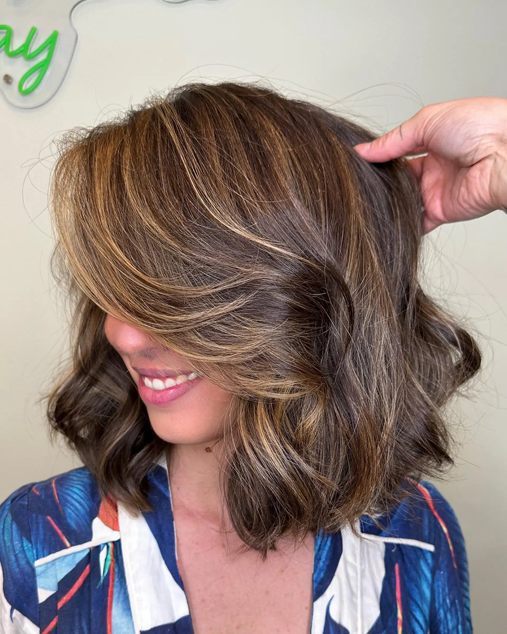 A textured shoulder-length brunette bob styled with soft waves and luminous caramel highlights. 1