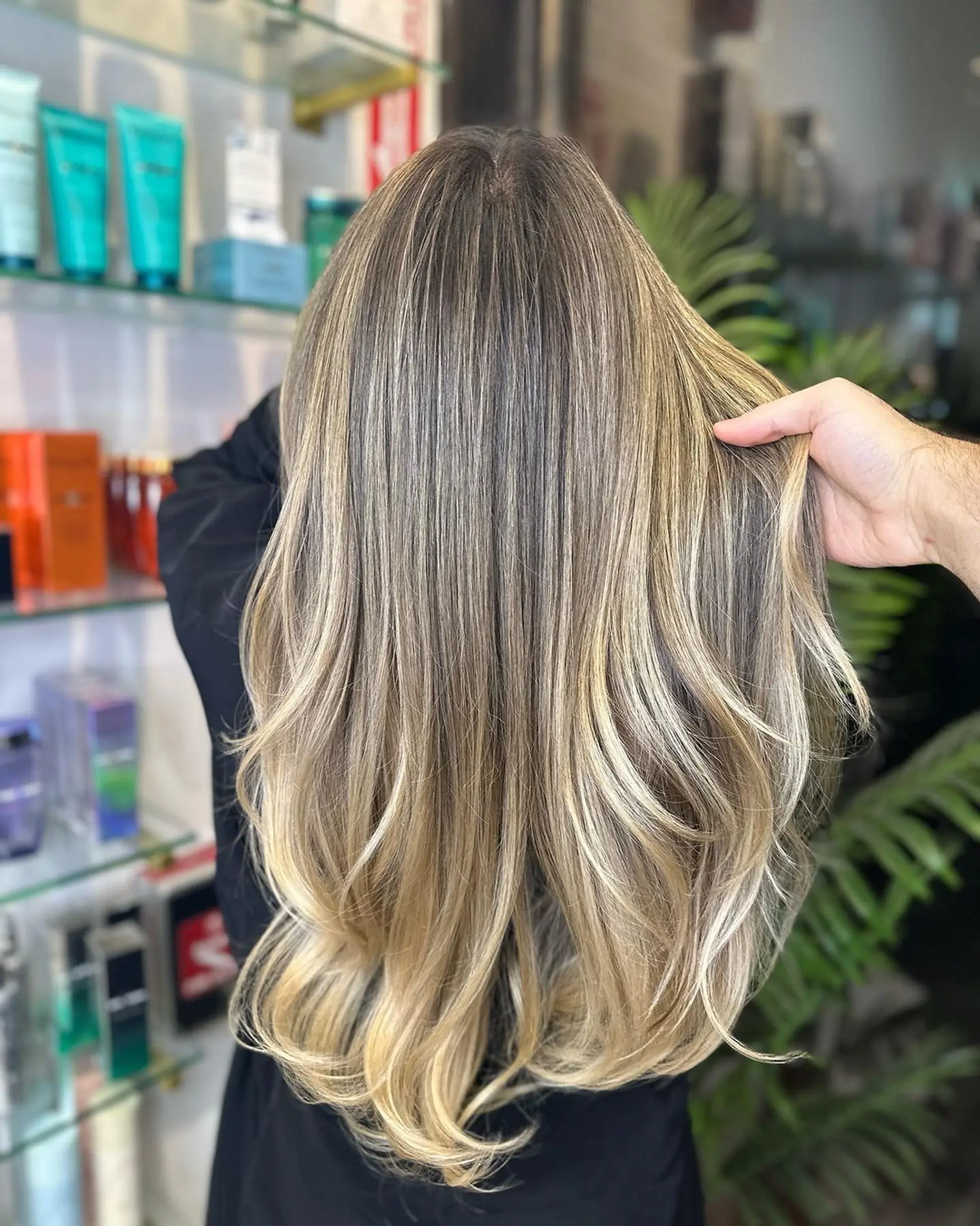 The hairstyle features very long, wavy hair with natural brunette roots transitioning into blonde through balayage. It has layered cuts for volume and soft waves for a beachy look. 1