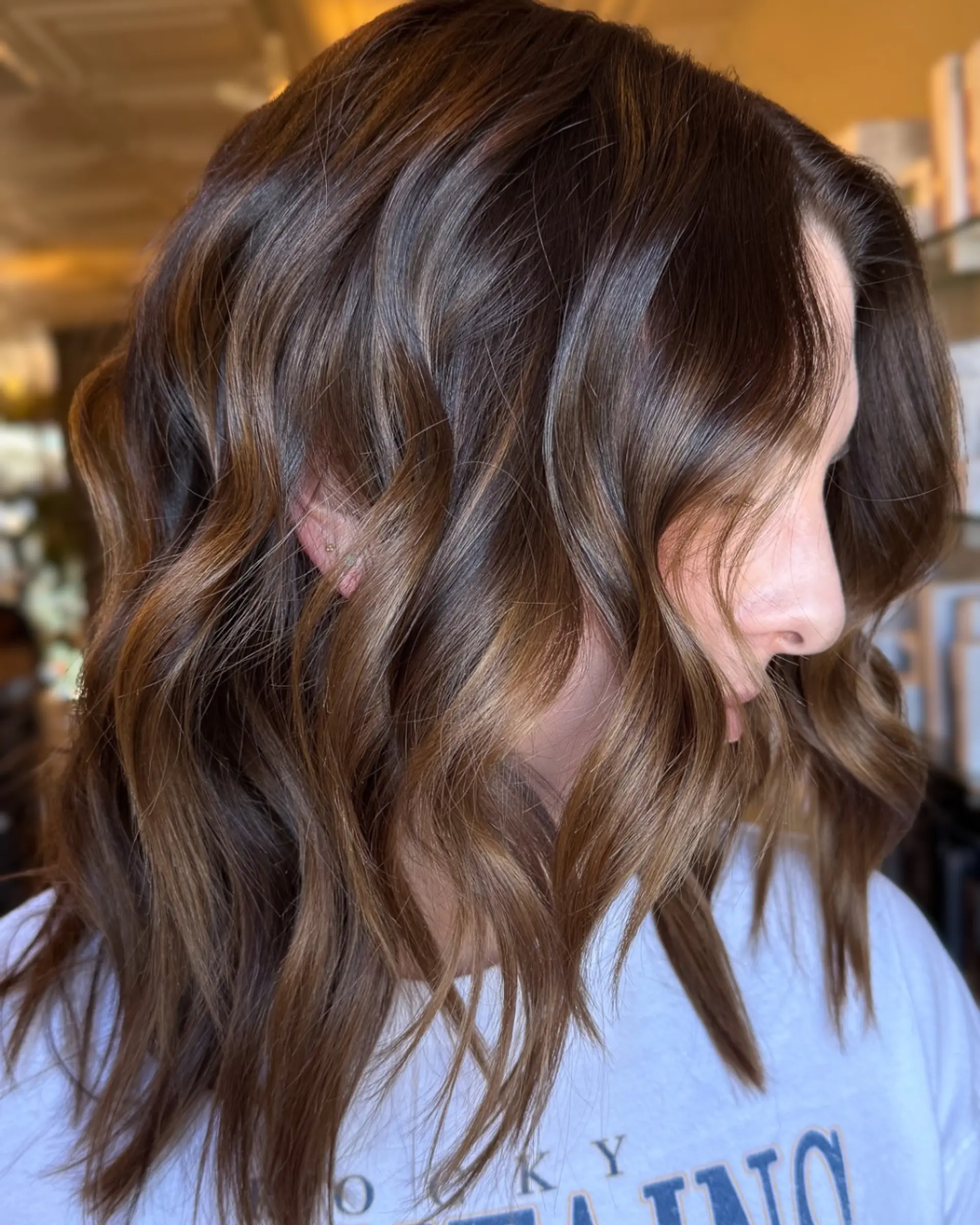 Medium-length rich brunette hair styled into soft, textured waves featuring seamless warm dimensional highlights. 1