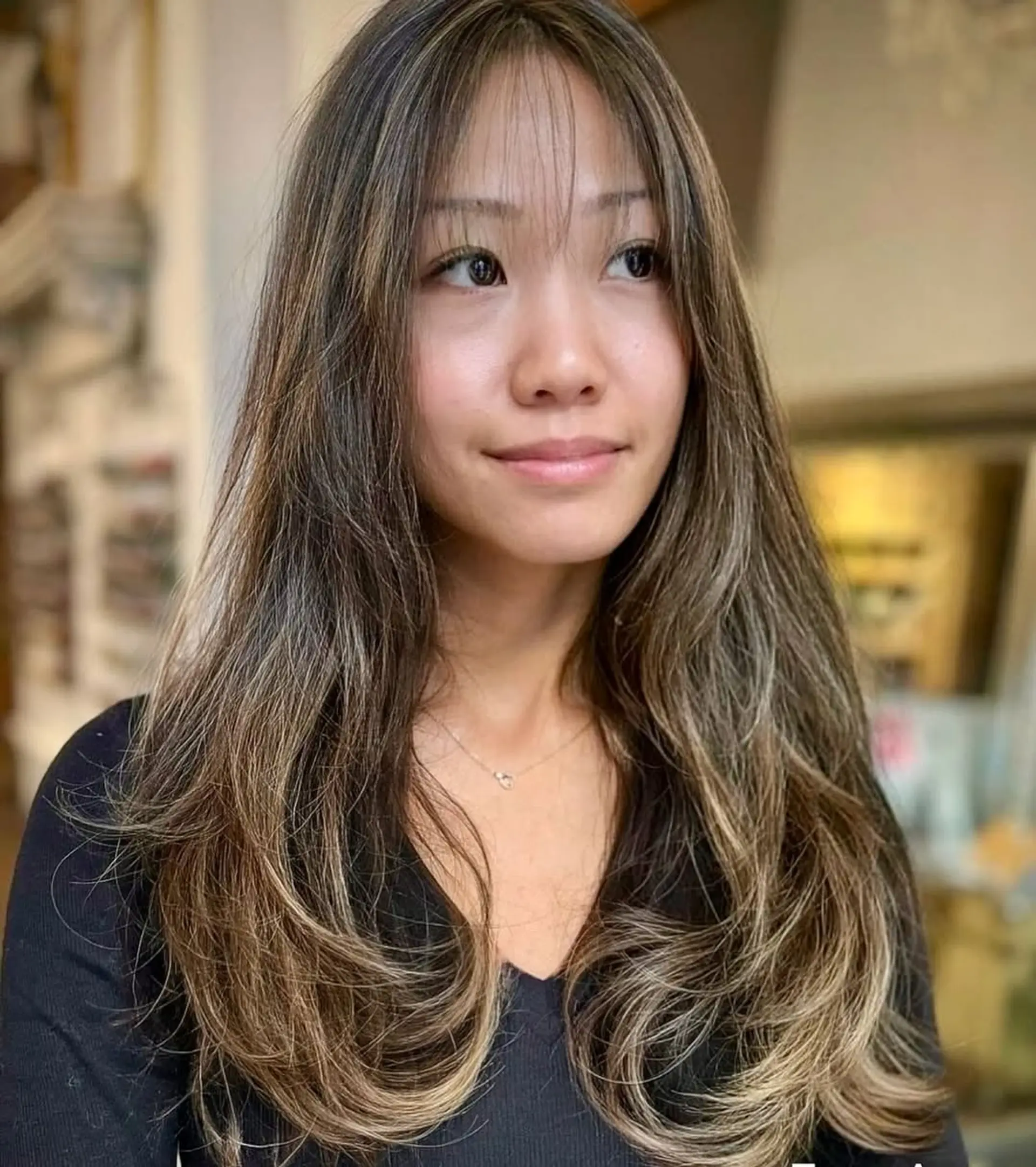 Long, wavy hair with face-framing layers and wispy bangs, enhanced by dark brown color and ash blonde highlights, featuring a subtle ombre effect. 1