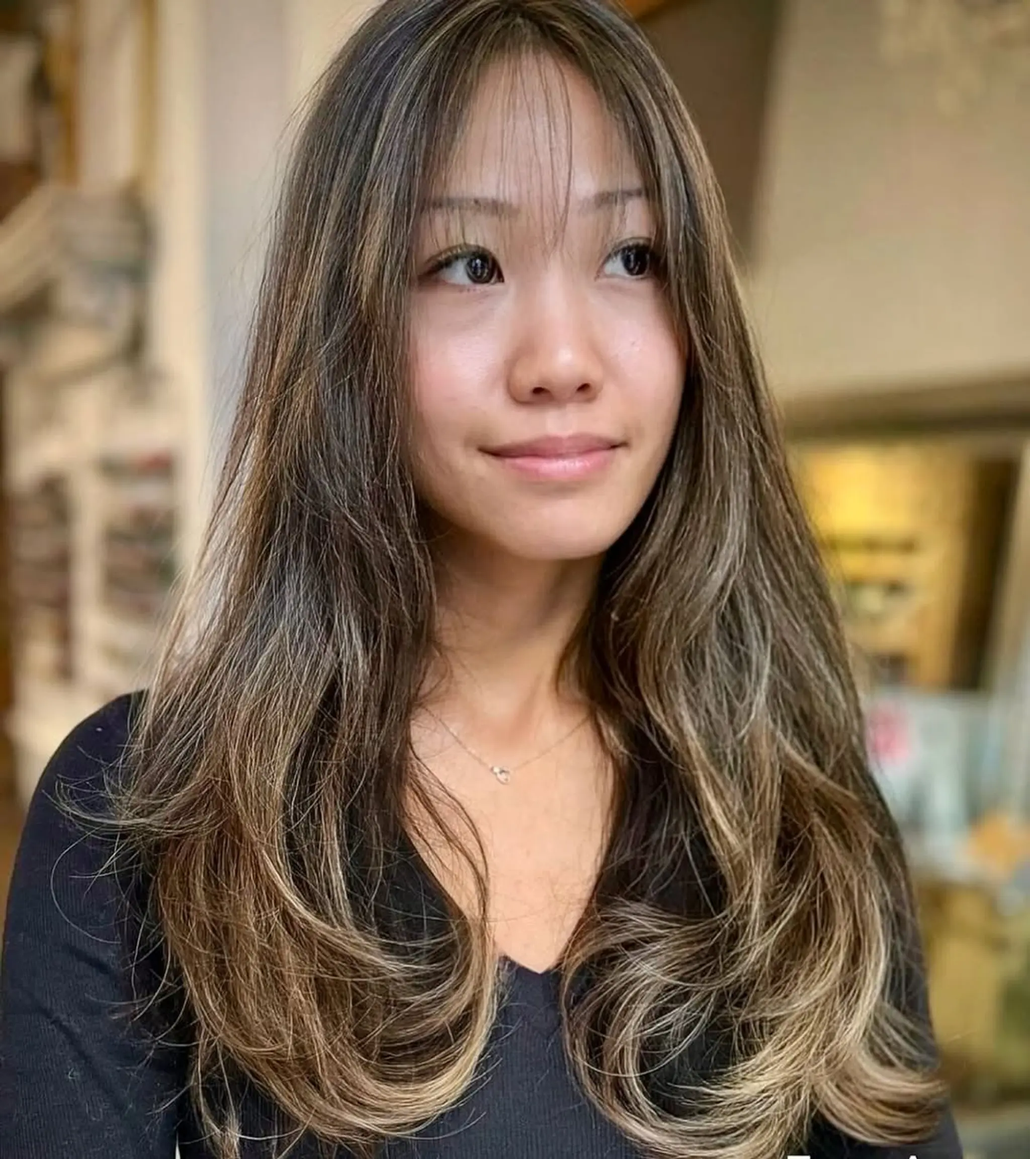 Long, wavy hair with face-framing layers and wispy bangs, enhanced by dark brown color and ash blonde highlights, featuring a subtle ombre effect.