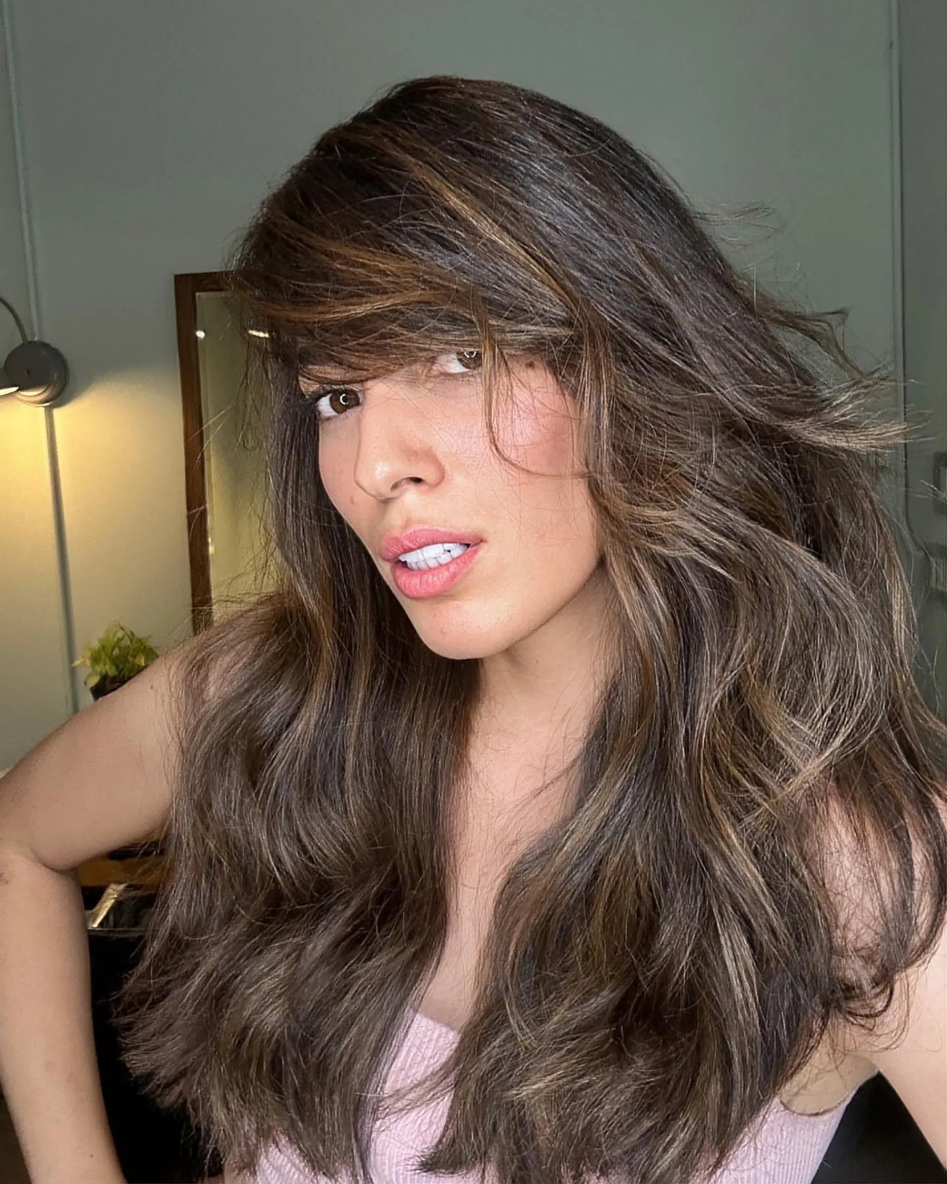 Voluminous long layered brunette hair featuring warm caramel highlights and face-framing side-swept bangs styled in soft waves. 1
