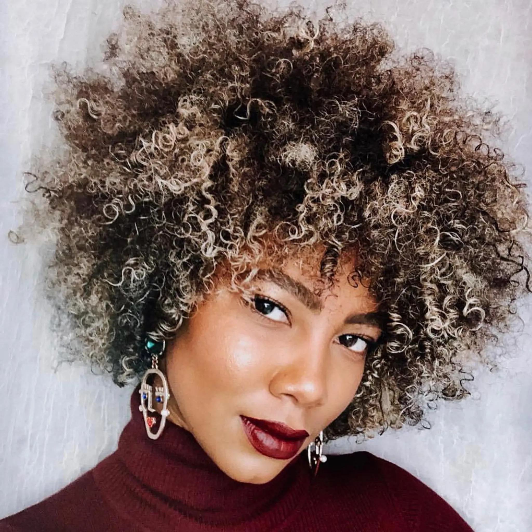 The hairstyle features a voluminous afro with defined and bouncy curls. The base color is black with ash blonde highlights throughout, giving the hair a frosted and modern look.