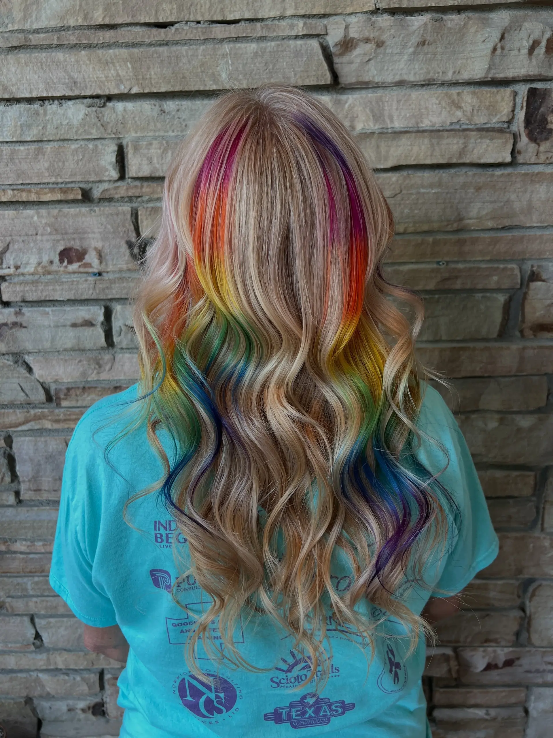 Long wavy blonde hair featuring vibrant rainbow peekaboo highlights layered underneath, styled in loose soft waves. 1