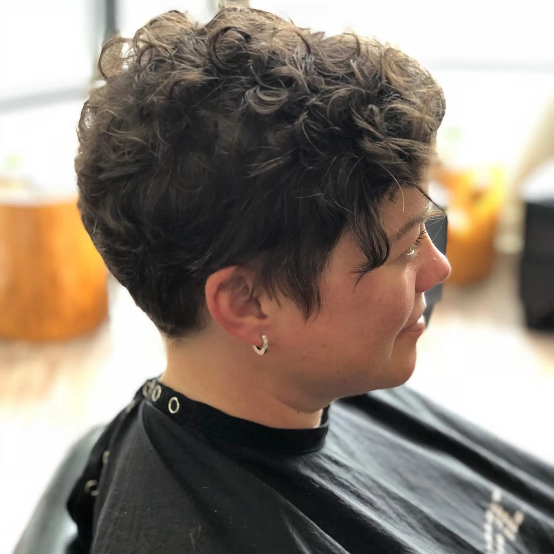 A short, tapered curly pixie haircut featuring a softly textured top and an asymmetrical fringe framing the face. 1