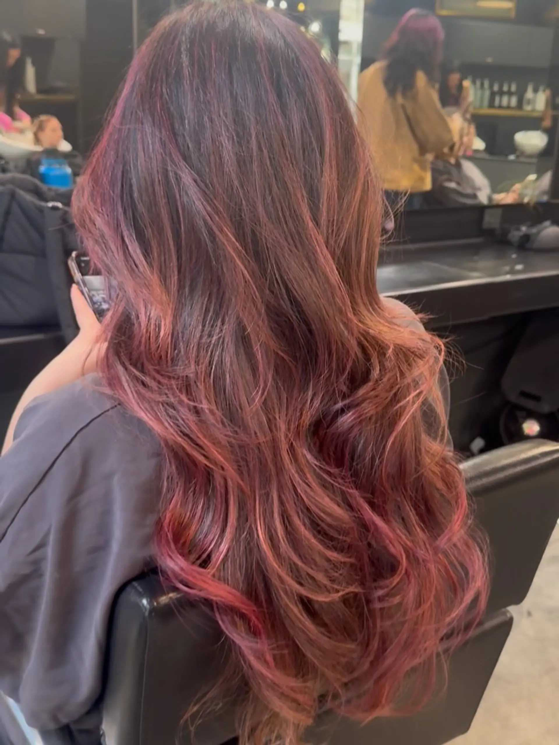 Long cascading layers on dark brunette hair infused with vibrant rose and red highlights, styled into loose bouncy waves. 1
