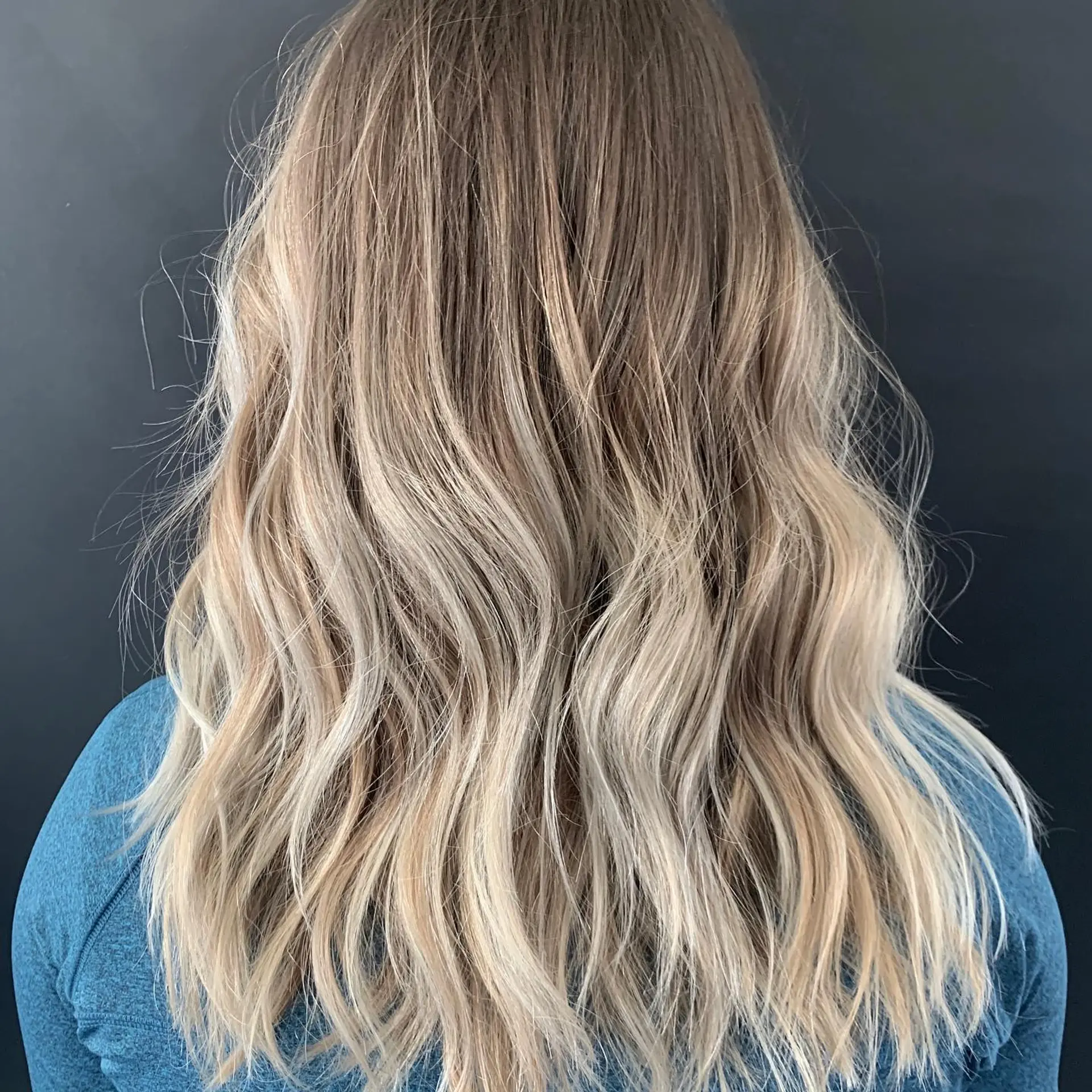 Long layered hair styled with relaxed, textured waves showcasing a dimensional blonde balayage with a soft root melt. 1