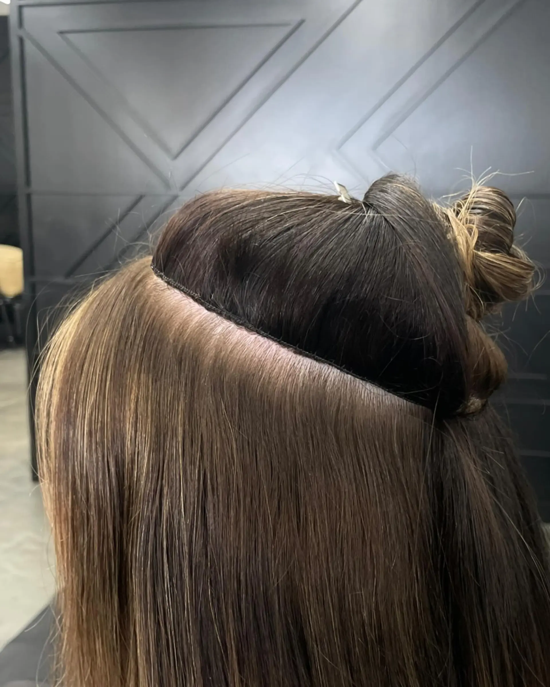 A close-up view displaying a seamless hand-tied extension track installed cleanly along a parted row of straight brunette hair. 1
