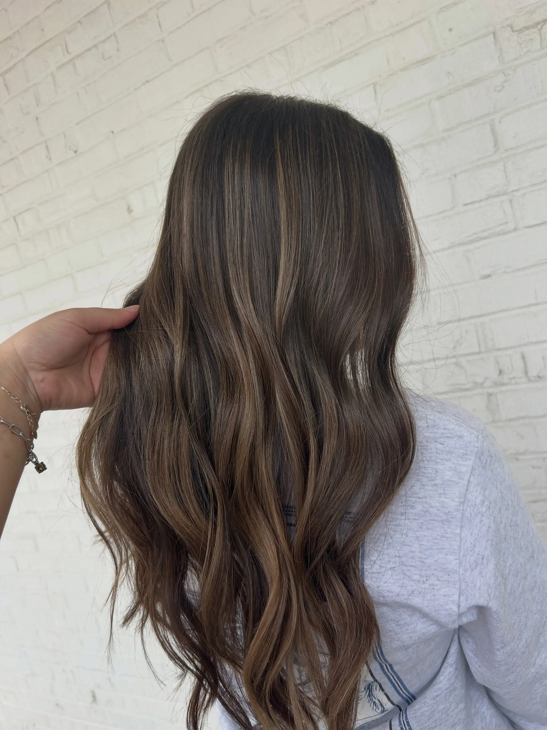 Long dark brown hair enhanced with subtle, lived-in light brown balayage highlights and styled in soft waves. 1