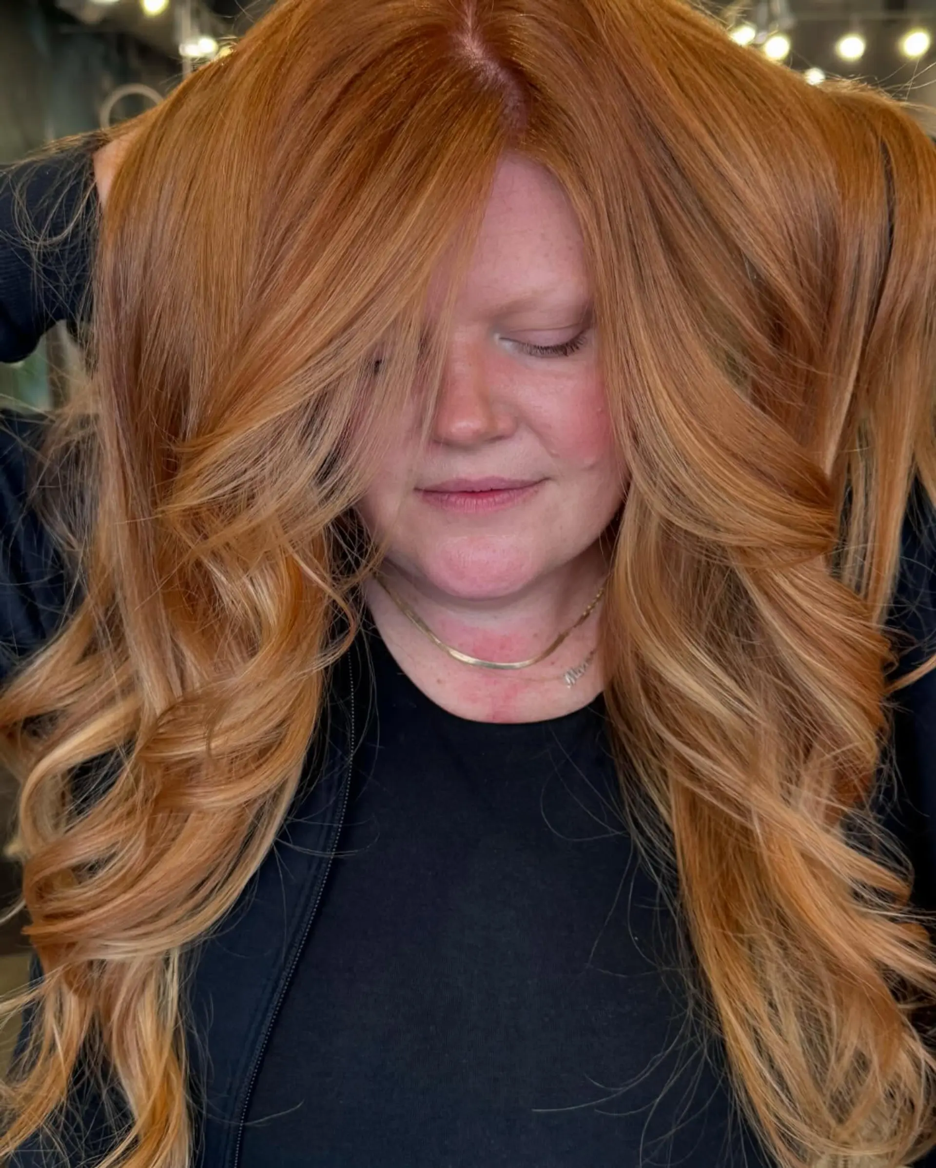 A long, wavy hairstyle with layered ends and face-framing layers, highlighted by a warm copper color with blonde balayage. 3