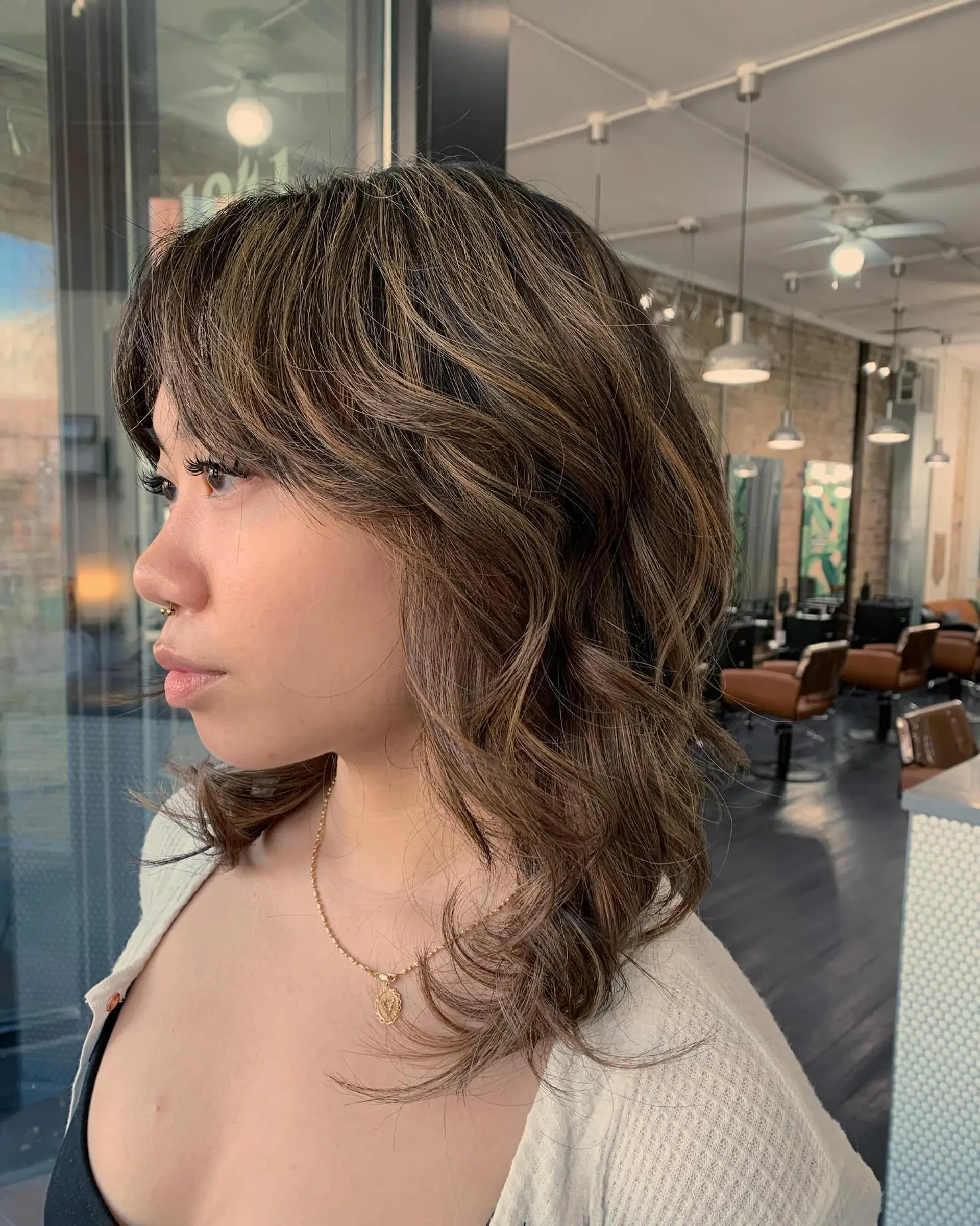 Medium-length haircut with face-framing curtain bangs, textured layers, and dimensional cool-toned milk tea highlights styled into soft waves. 1