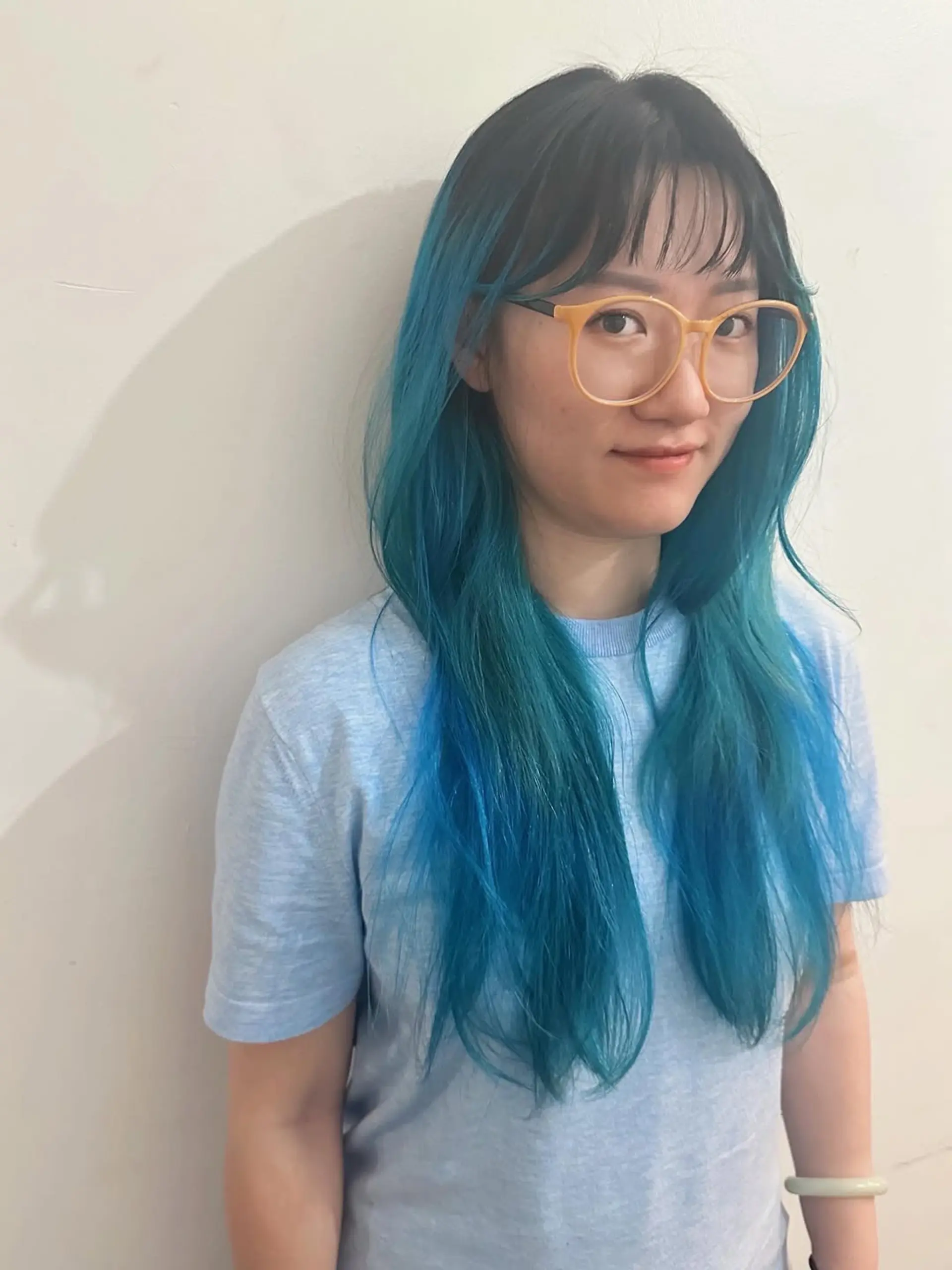 This captivating hairstyle features long, straight hair with a striking teal and blue ombre effect, graduating from dark natural roots. The vibrant color adds a bold and trendy statement, complemented by chic, blunt bangs that softly frame the face. 1