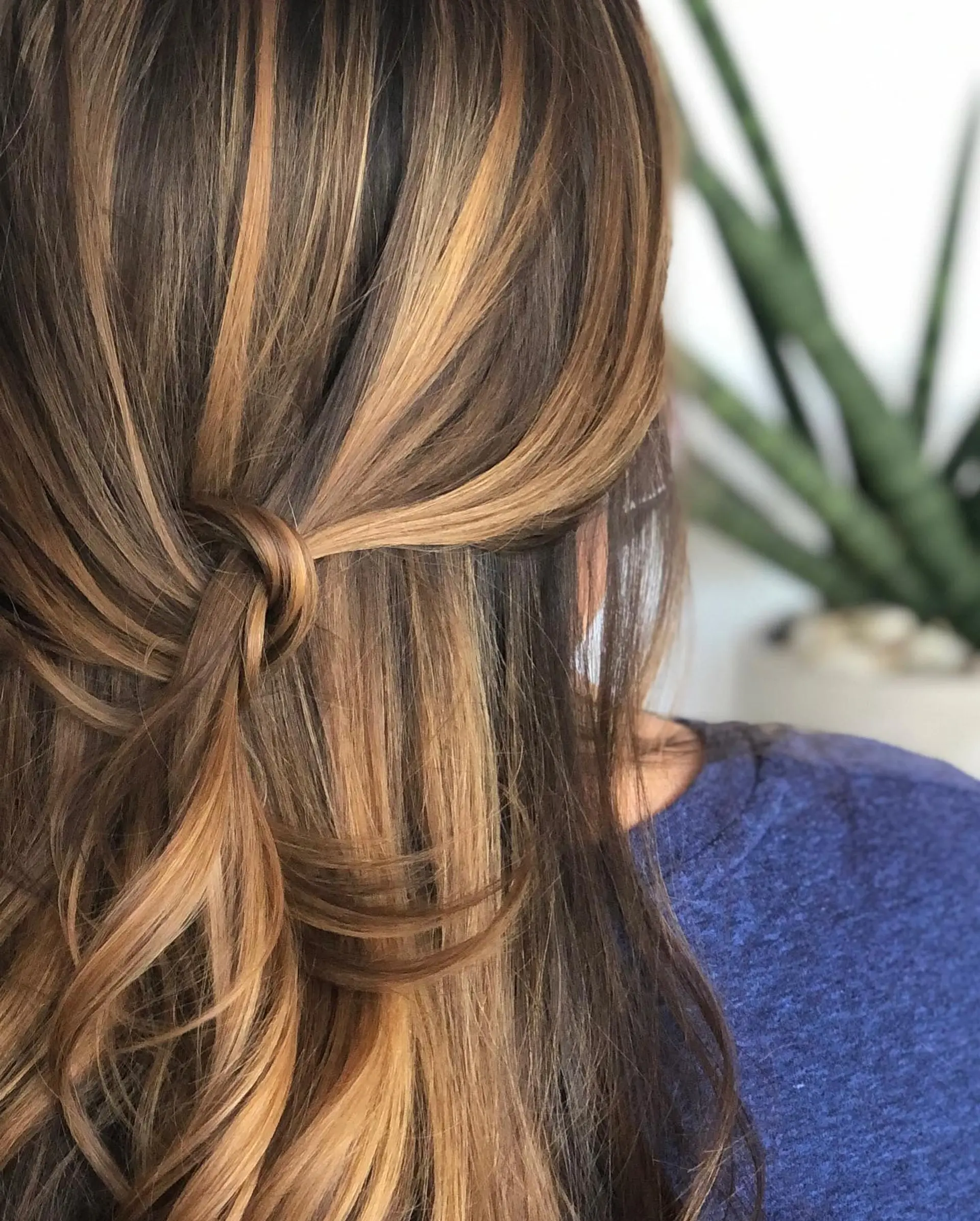 Long dimensional brunette hair with warm honey-caramel balayage, gathered into an effortless half-up knotted accent. 1