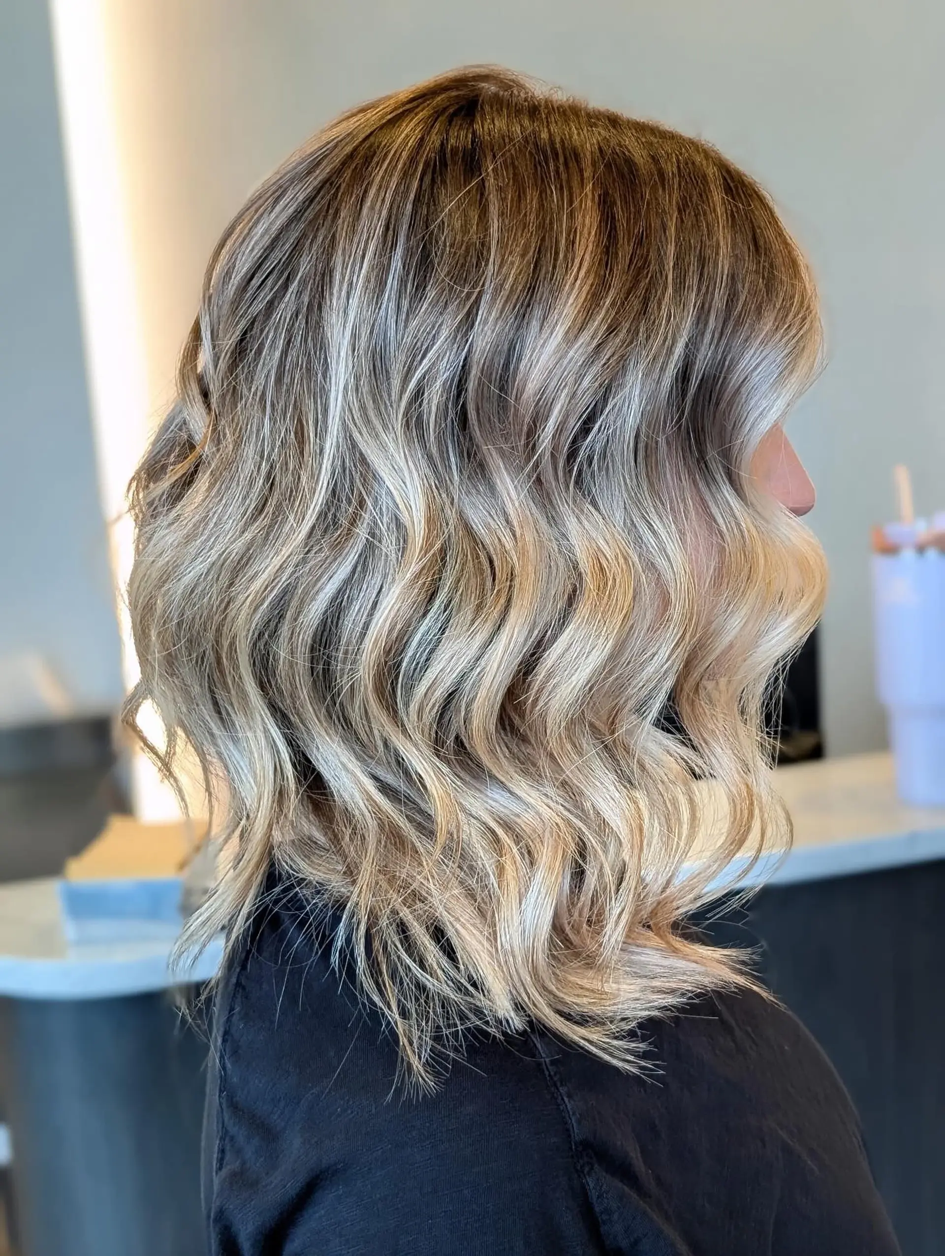 A textured, shoulder-skimming lob featuring cool-toned ash blonde foilyage highlights over a natural brown base, styled with soft beach waves. 1