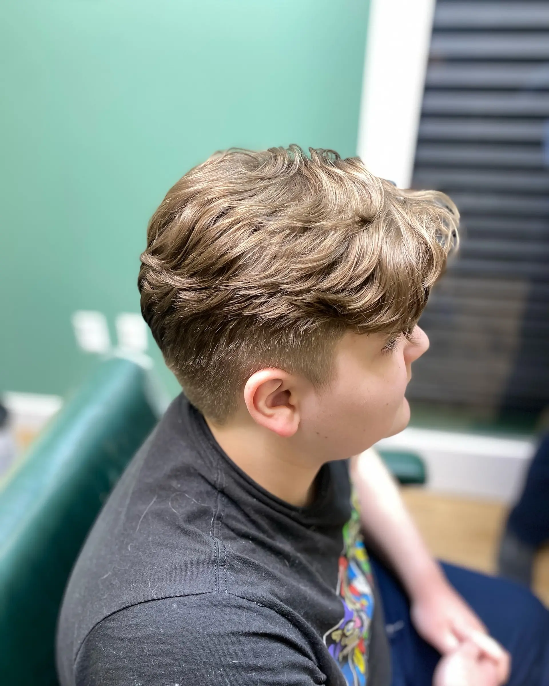 A modern short haircut featuring clipper-tapered sides and a textured, naturally wavy top styled forward. 1