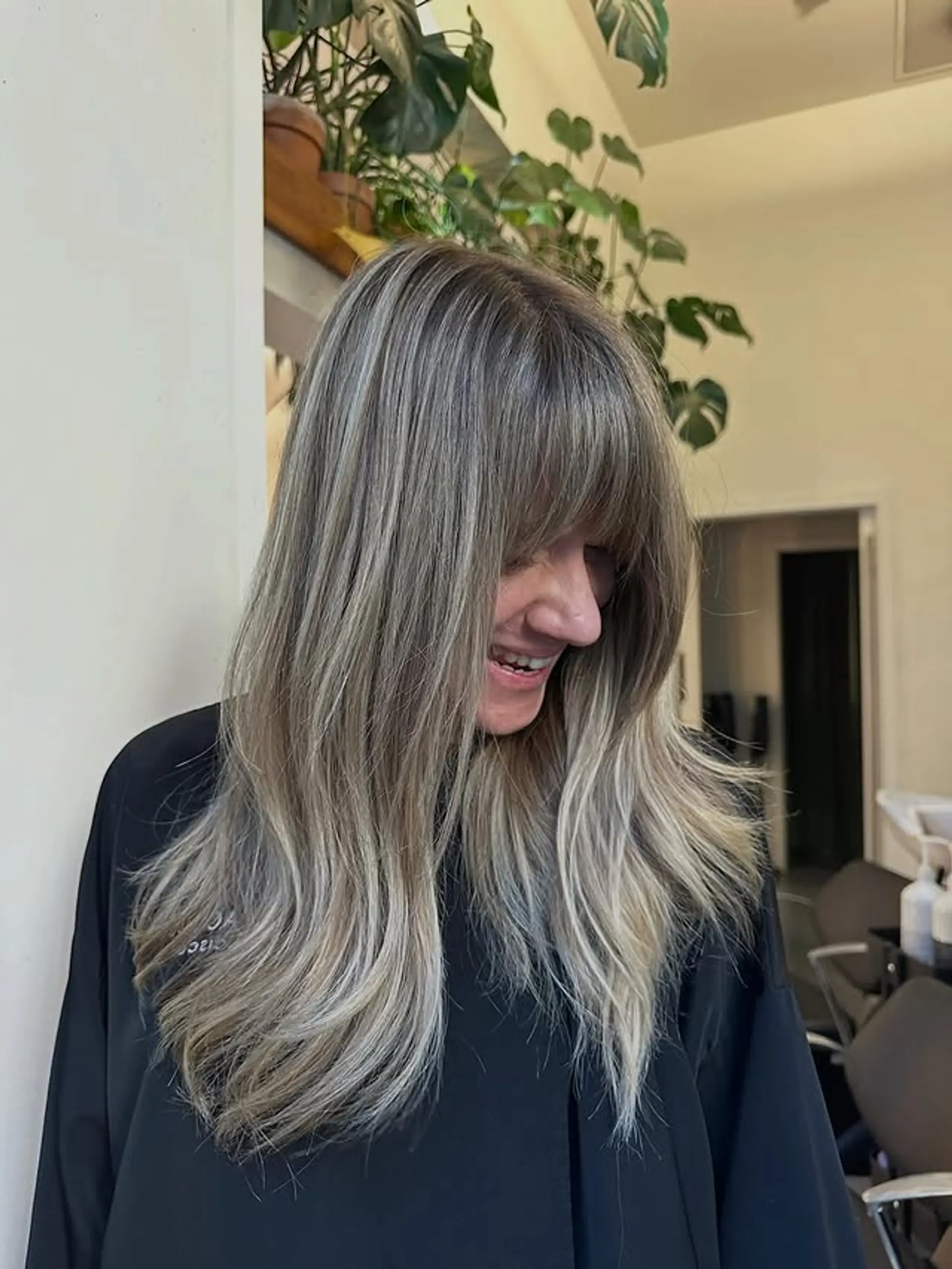 A long, layered hairstyle with a full fringe, soft waves, and face-framing layers. The hair is styled with an ash blonde balayage. 1