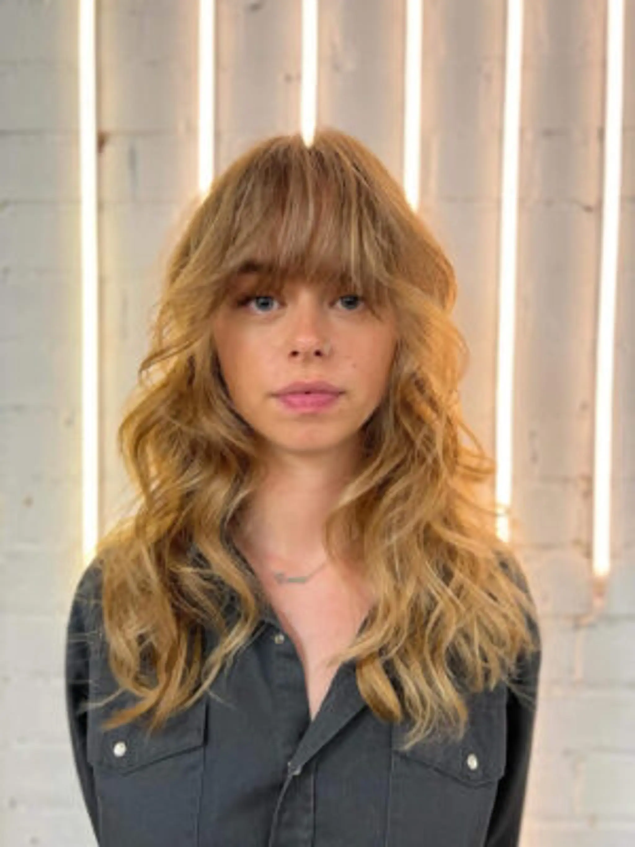 Medium-length honey blonde waves with layered texture and a full fringe providing a soft, tousled look.