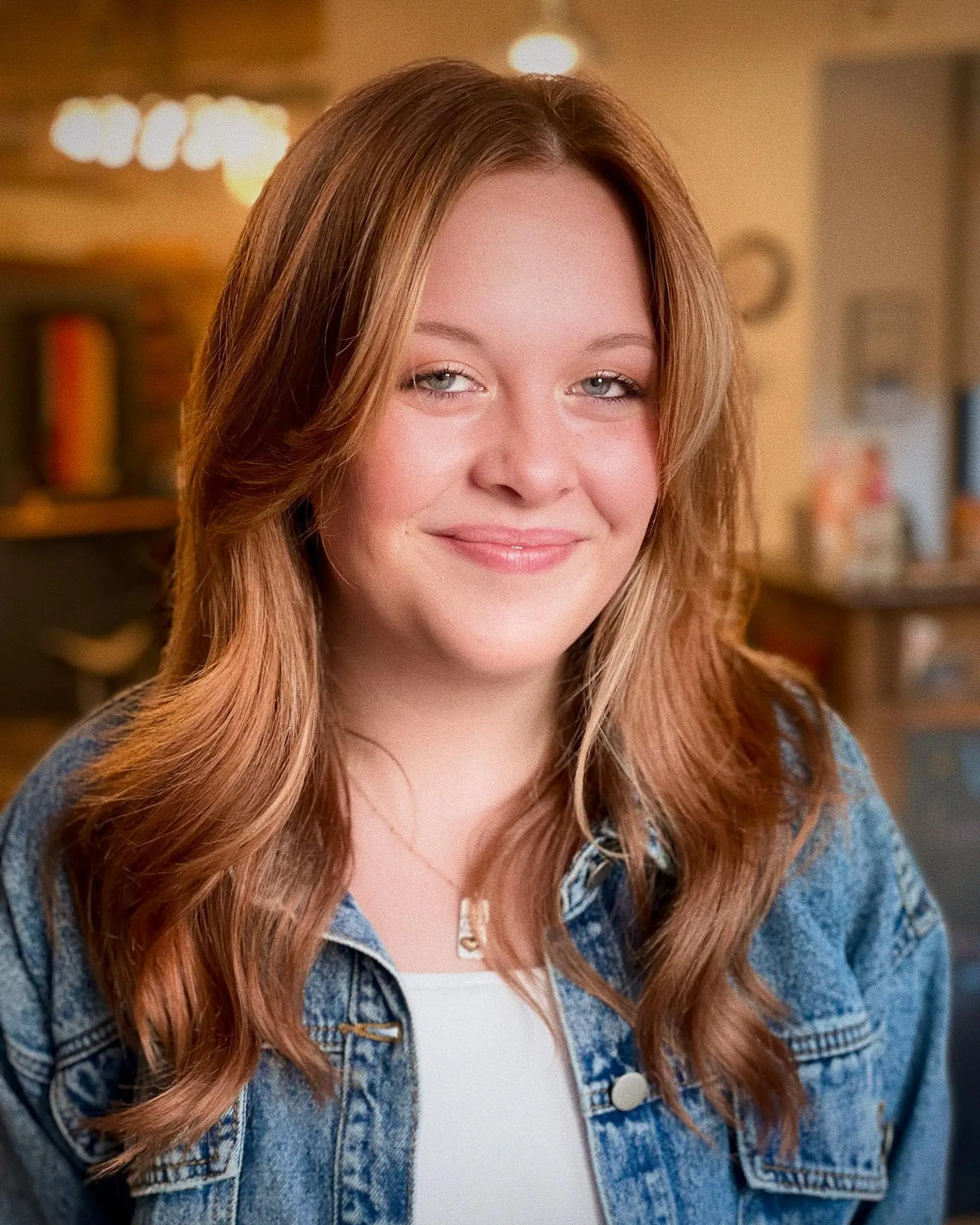 Medium-length layered haircut with soft curtain bangs and gentle waves in a warm strawberry blonde shade. 1