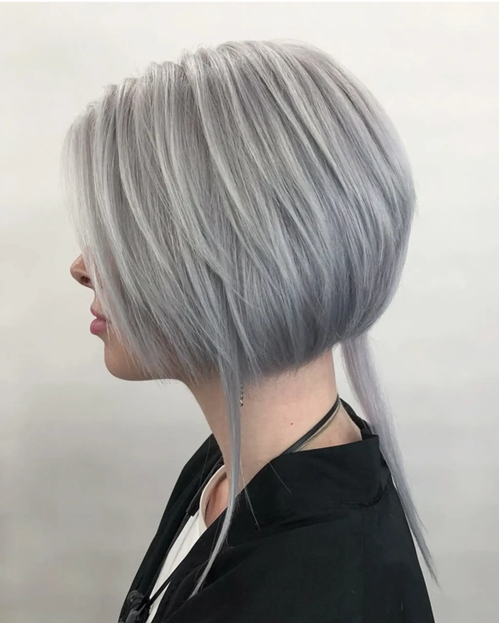 An edgy and modern asymmetrical bob with an A-line shape, shorter at the back and longer towards the front. The front side strands are significantly longer, and the hair is straight and layered for texture and movement, finished with a striking silver and platinum color. Includes a side-swept bang for an added sophisticated touch.