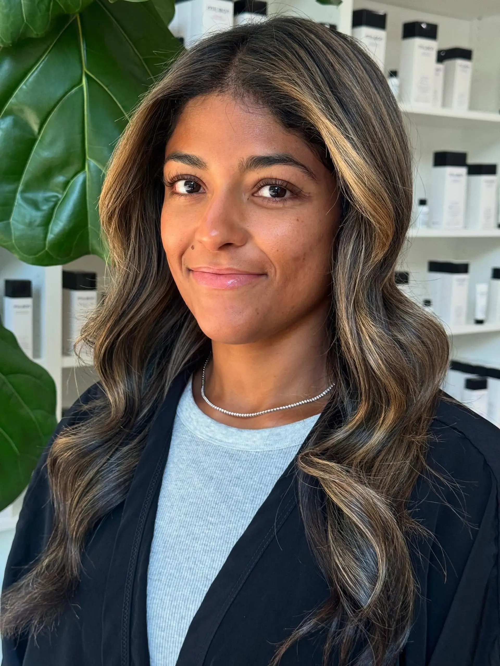 Long dark brown hair highlighted with warm caramel and golden blonde balayage, styled in soft, bouncy waves with face-framing layers. 1