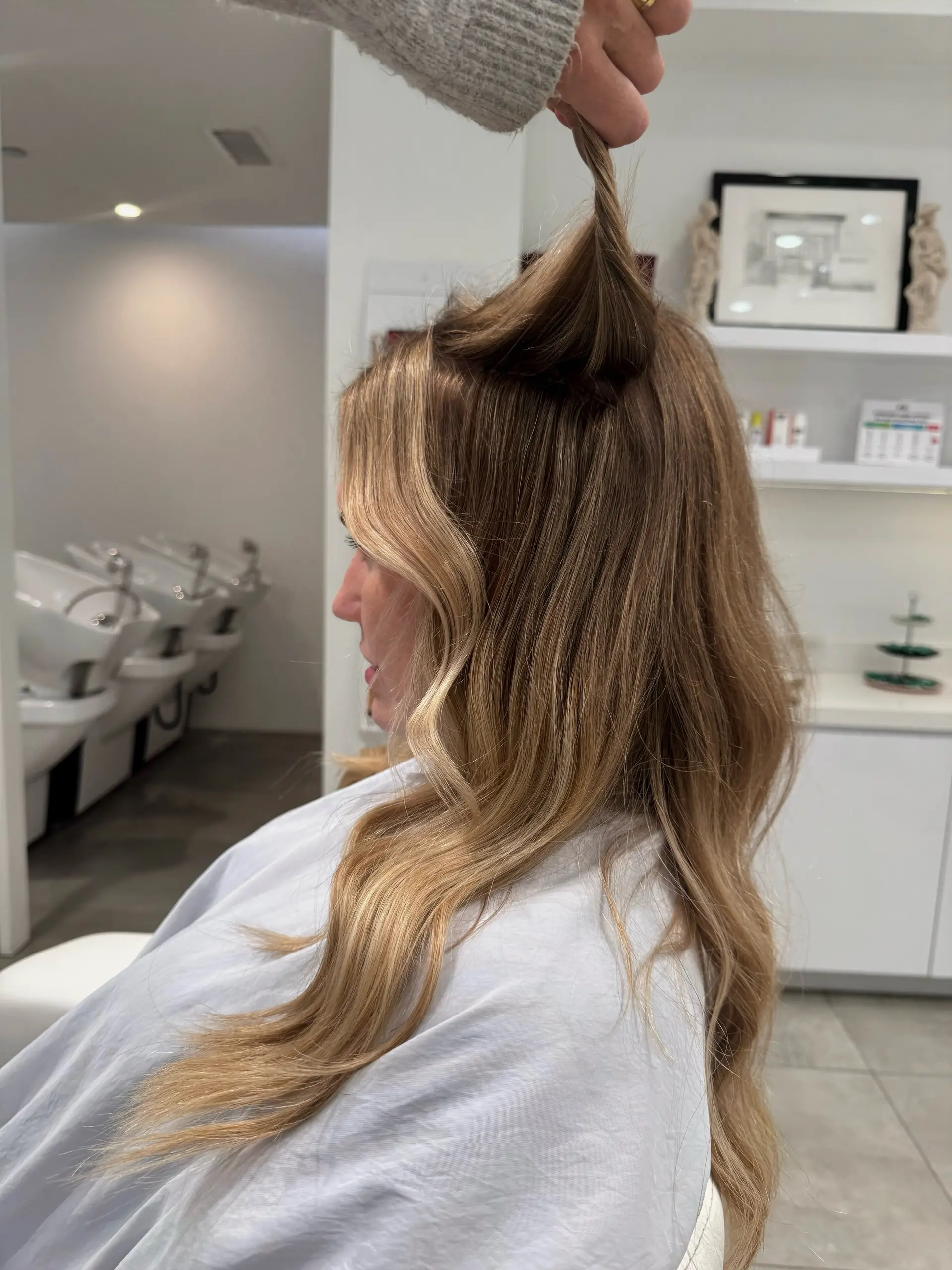 Long honey blonde hair styled in soft, loose waves, with a section gently lifted to showcase seamless, invisible extension placement. 1
