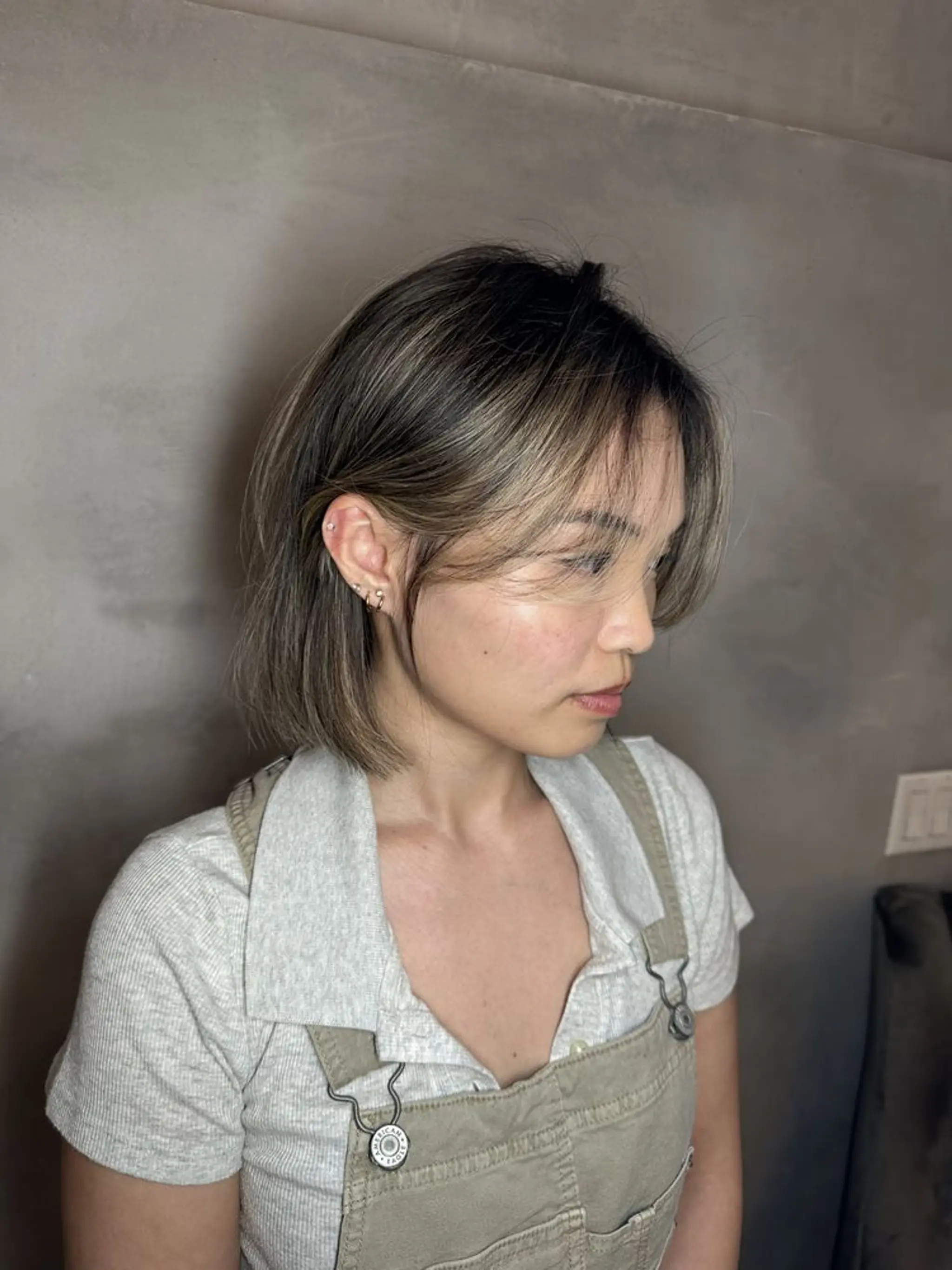 A medium-length ash brown bob with natural black undertones, featuring straight hair with minimal layers and side bangs that frame the face.