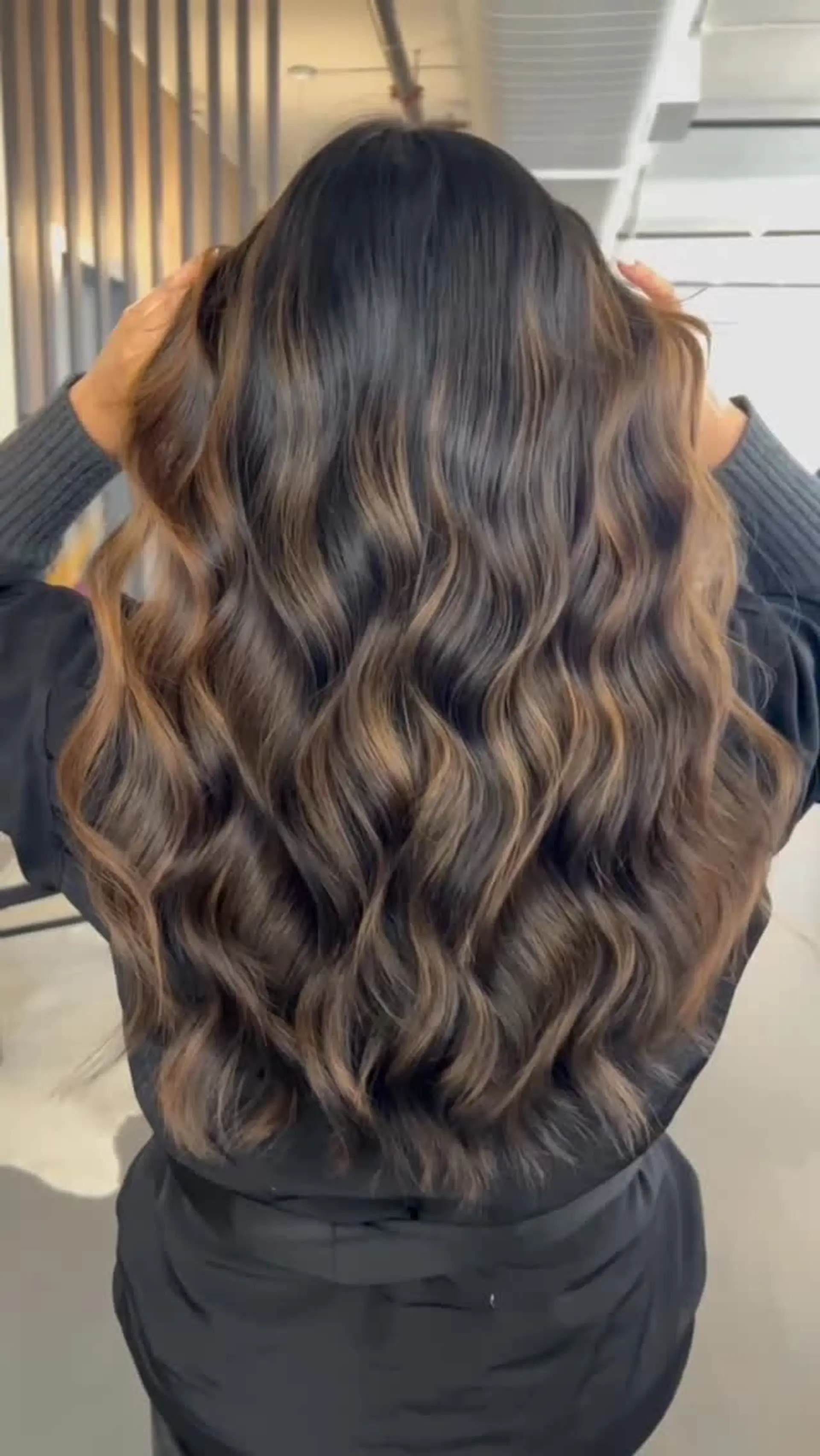 Long dark brown hair styled in soft body waves with seamless caramel balayage highlights. 1