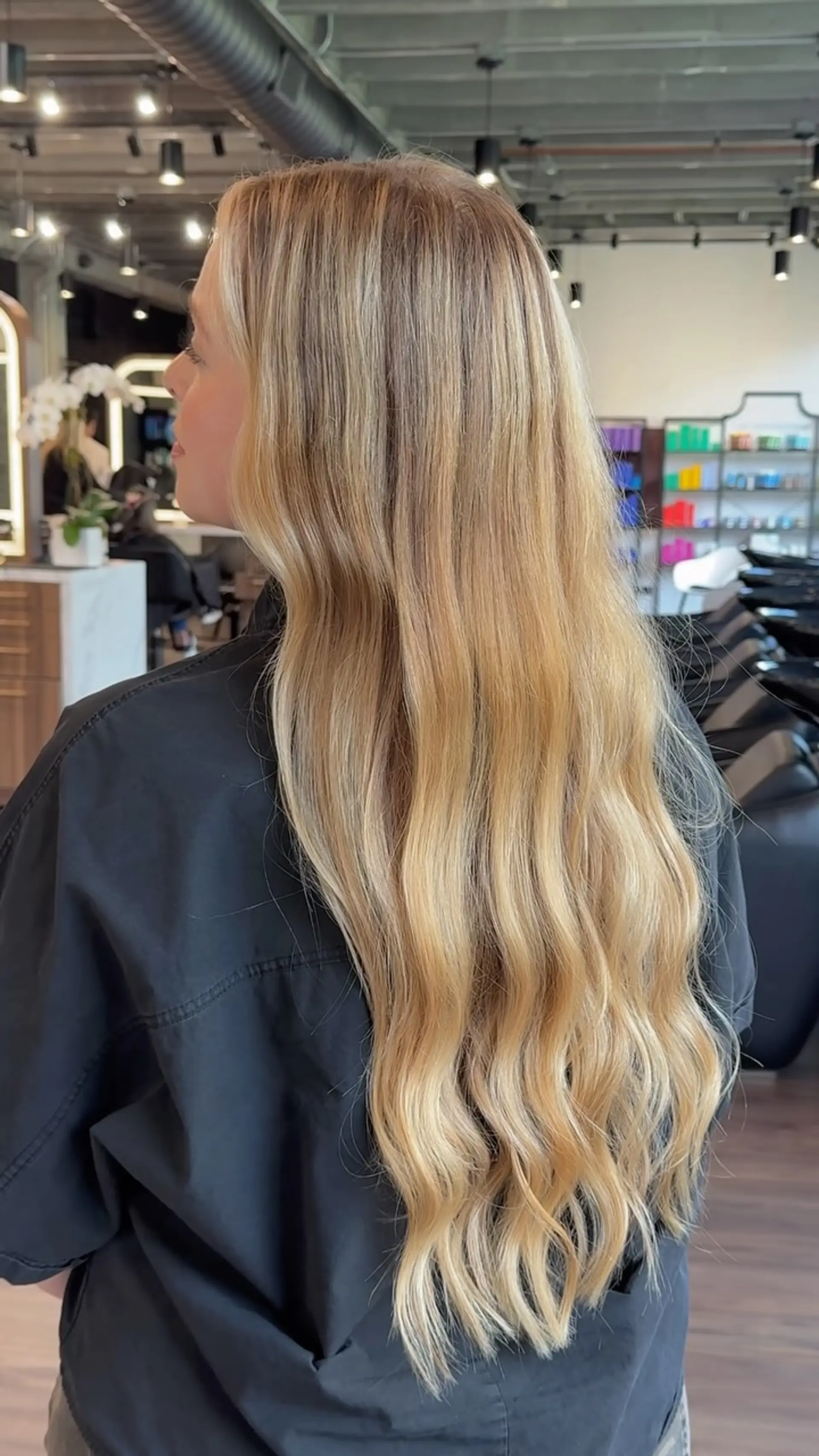 Long honey blonde hair styled with subtle, natural-looking body waves cascading down the back. 1