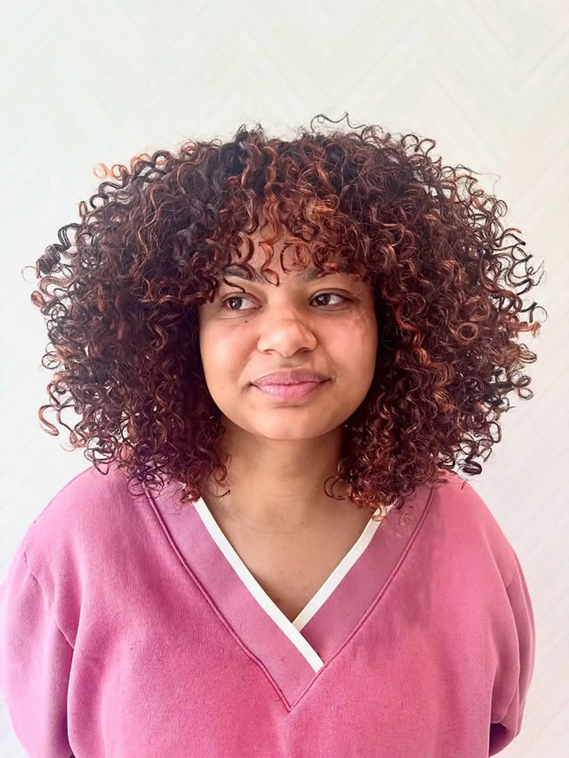 A medium-length curly hairstyle with copper tones and bangs. 1