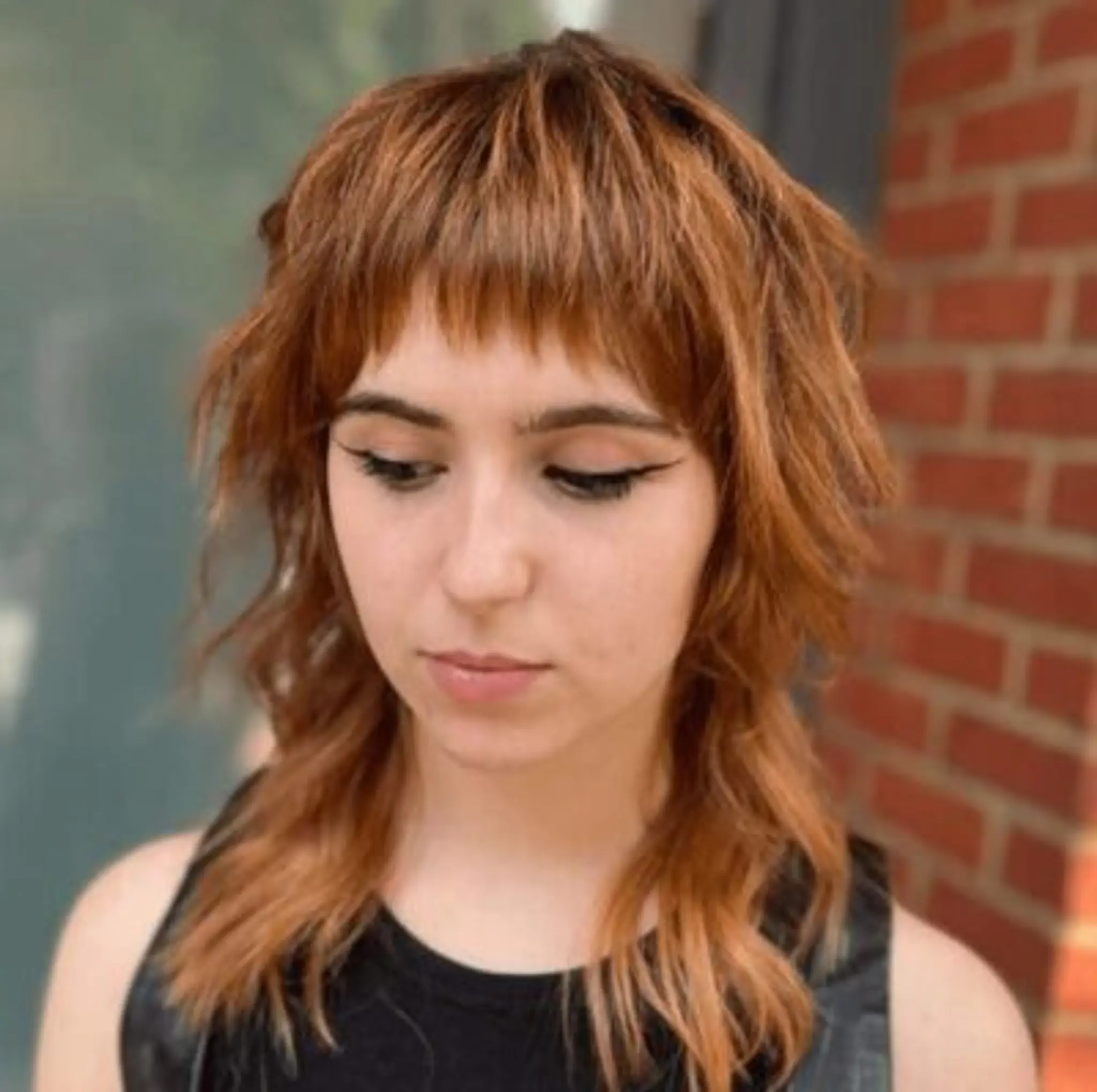 An edgy shag cut with a short, blunt fringe, medium length, and tousled wavy texture in a vibrant copper red hue.