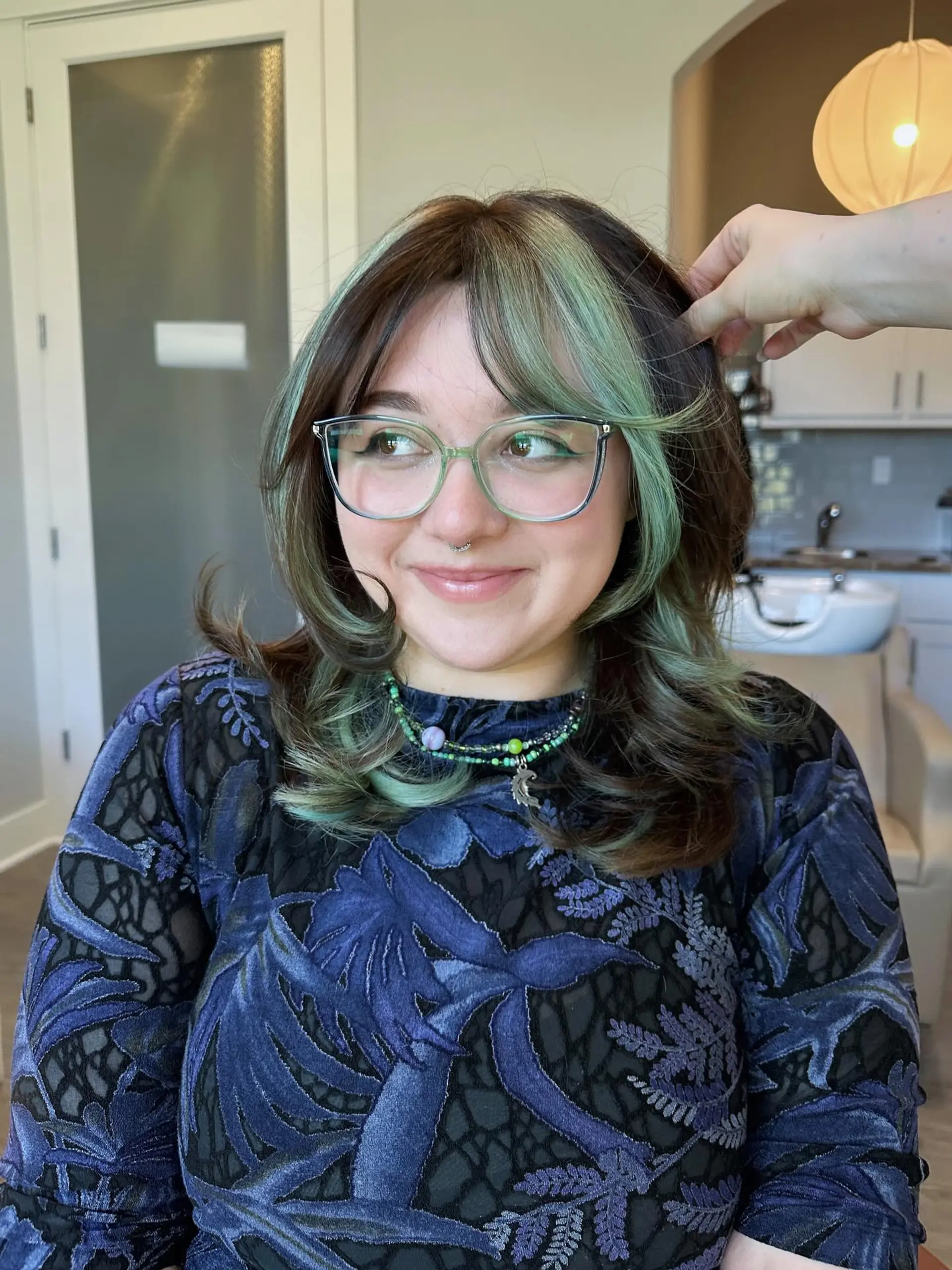 A trendy medium-length layered shag with voluminous, face-framing curtain bangs. The style features a bold 'money piece' highlight in vivid light green, contrasting sharply with the dark brown base color, extending through the ends in a wavy texture. 1