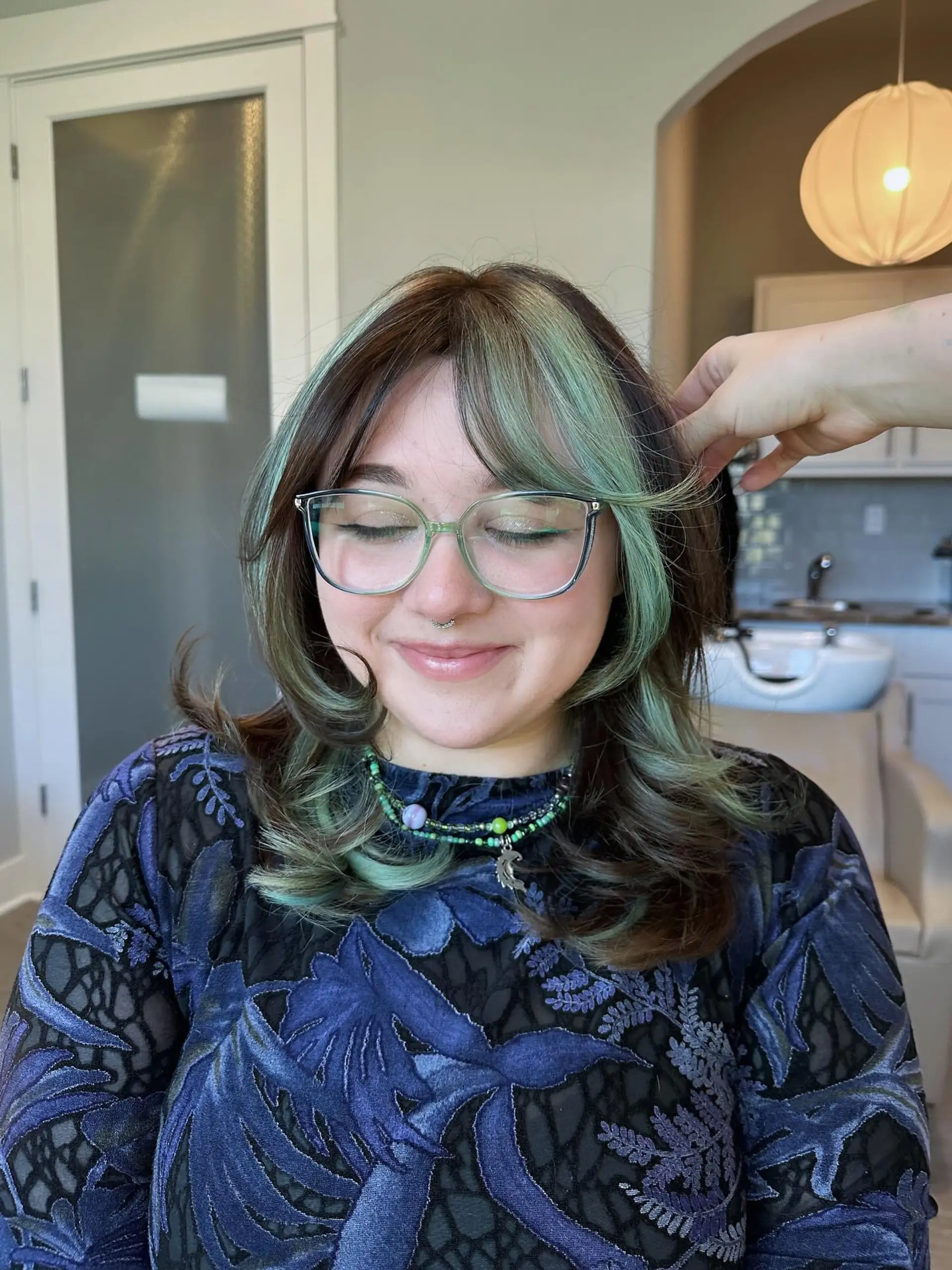 A trendy medium-length layered shag with voluminous, face-framing curtain bangs. The style features a bold 'money piece' highlight in vivid light green, contrasting sharply with the dark brown base color, extending through the ends in a wavy texture. 2