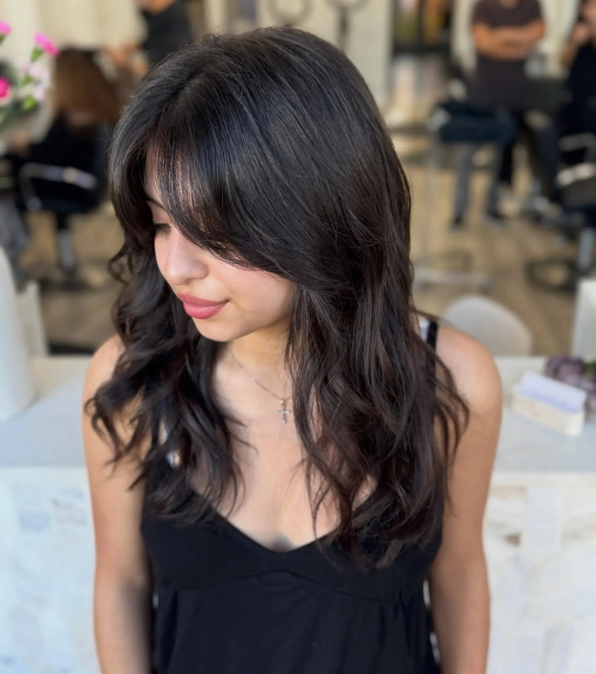 Long dark brown hair cut with soft cascading layers and wispy, face-framing curtain bangs styled into gentle loose waves. 1