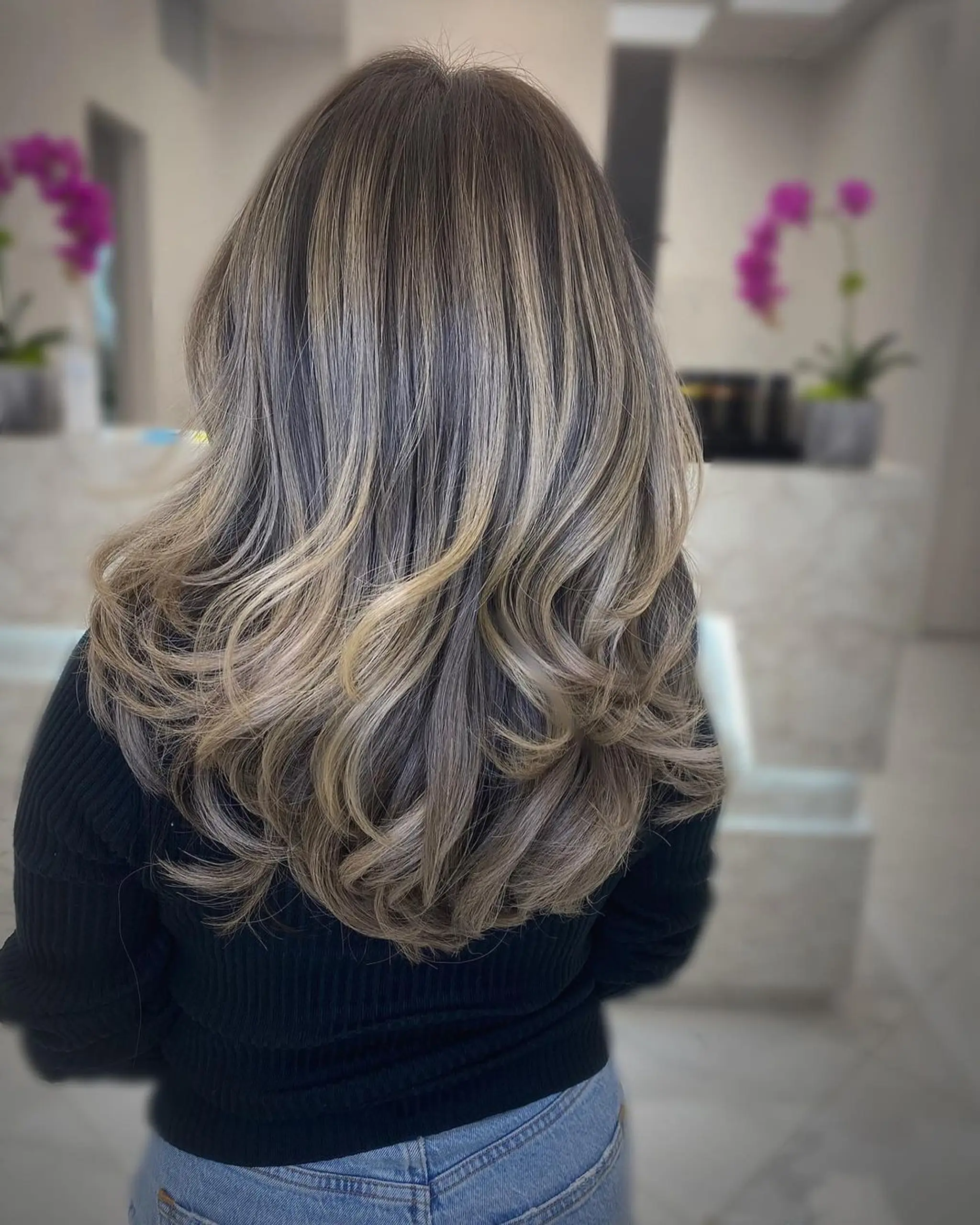 A long wavy hairstyle with a blend of blonde, brown, and ash blonde hues using a balayage technique. It features soft waves and a layered cut for added texture and volume.