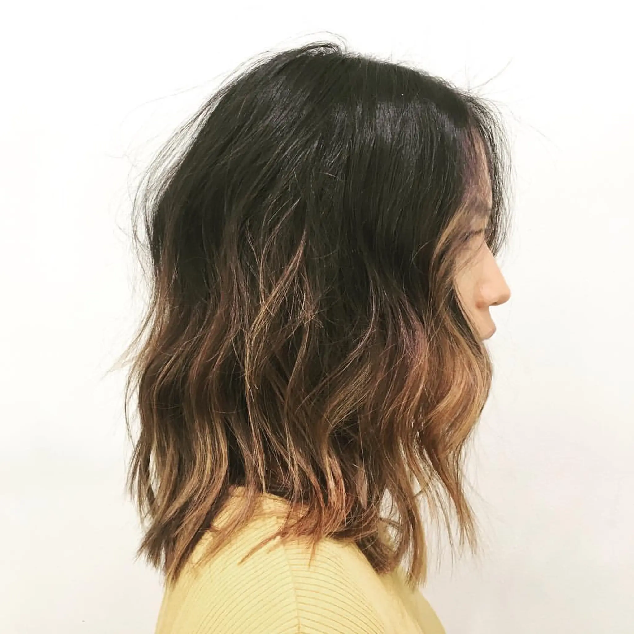 Medium-length wavy hairstyle with black roots transitioning to brown and blonde. Tousled waves, layered cut, and textured for volume.