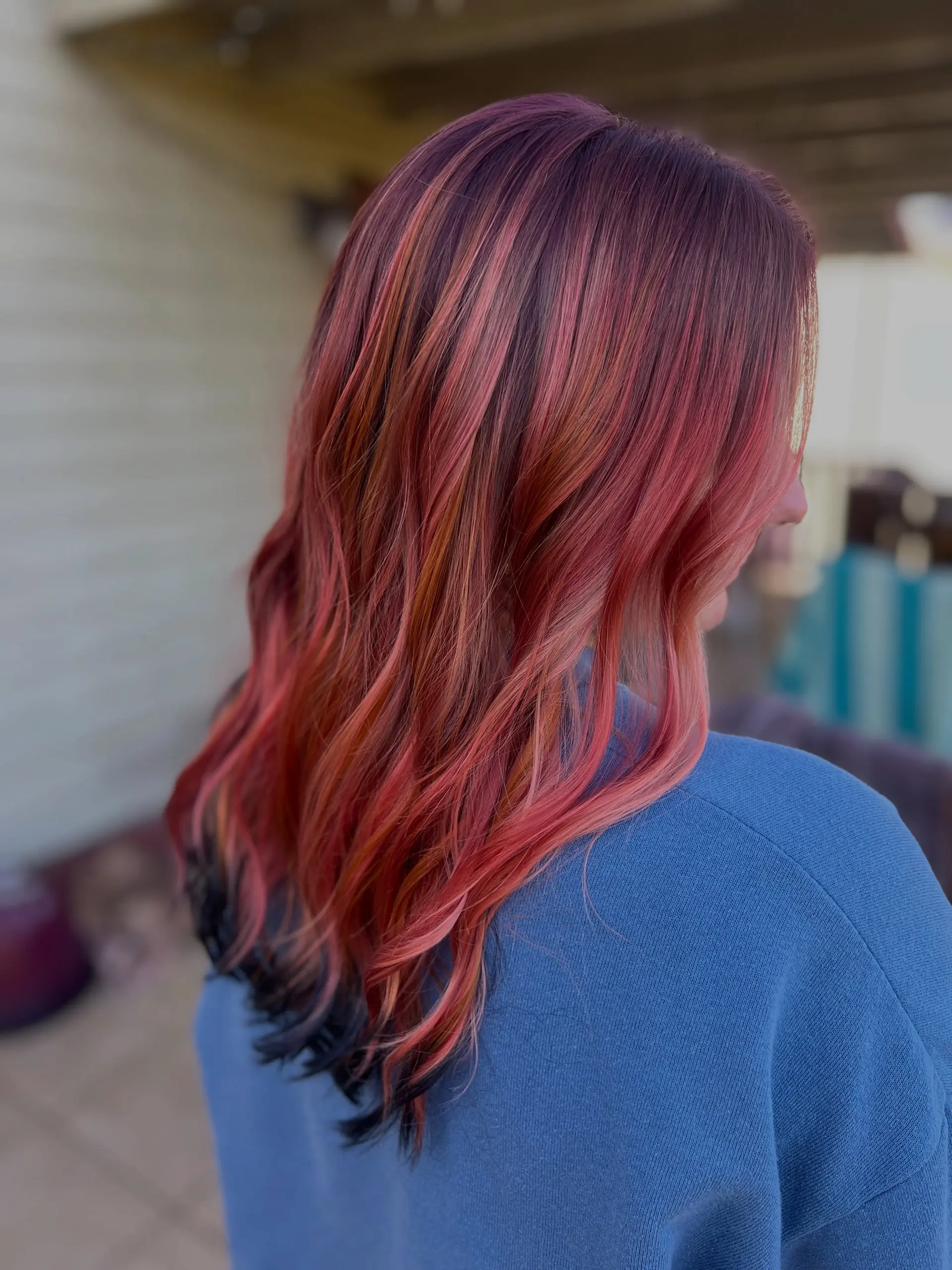 Long hair styled in soft waves showcasing a vibrant color melt of pink, coral, and copper highlights over deep roots with dark tips. 1