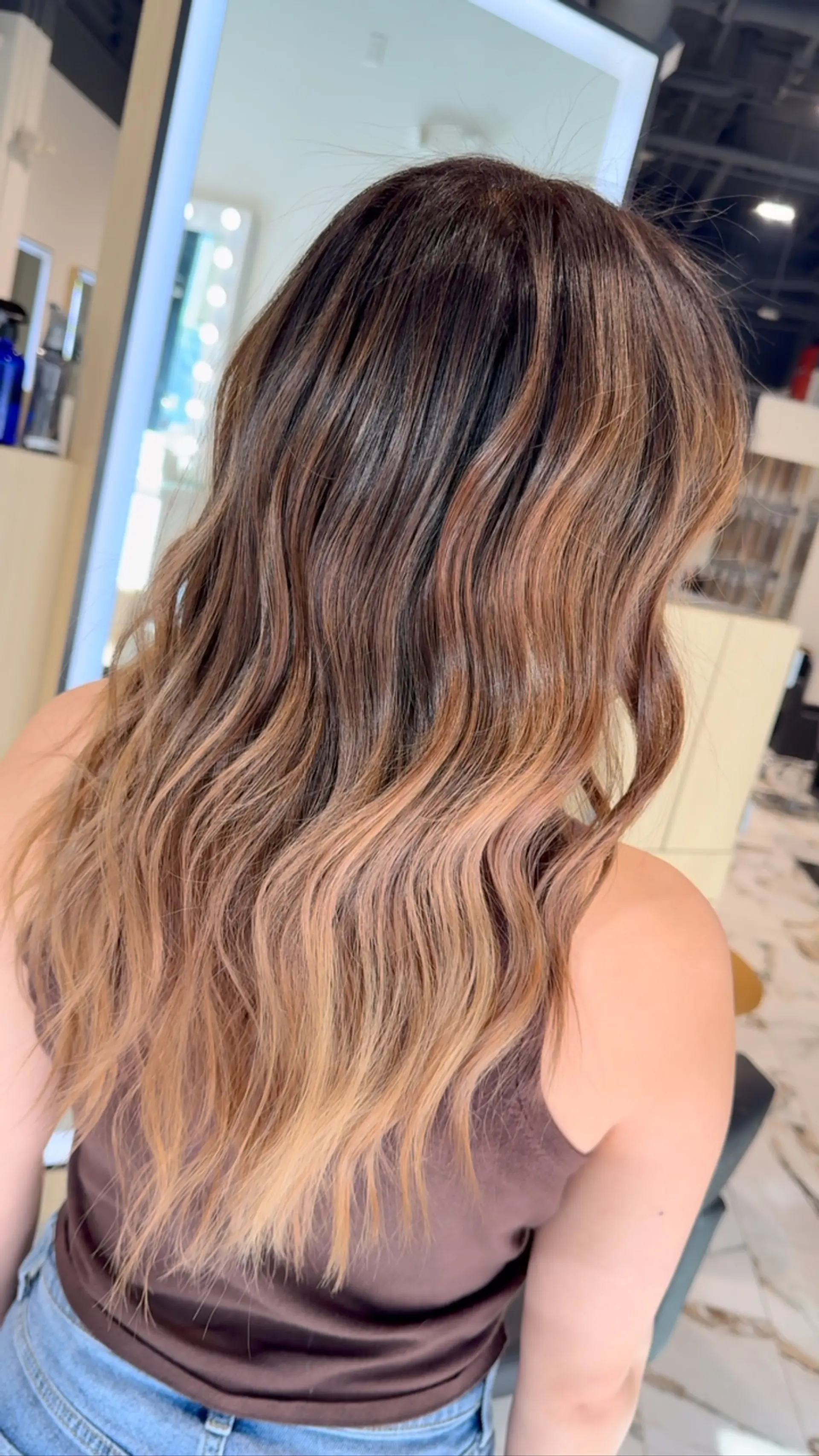 Long wavy hair featuring a seamless bronde highlight blend transitioning from a rich brunette root into warm caramel and honey tones. 1