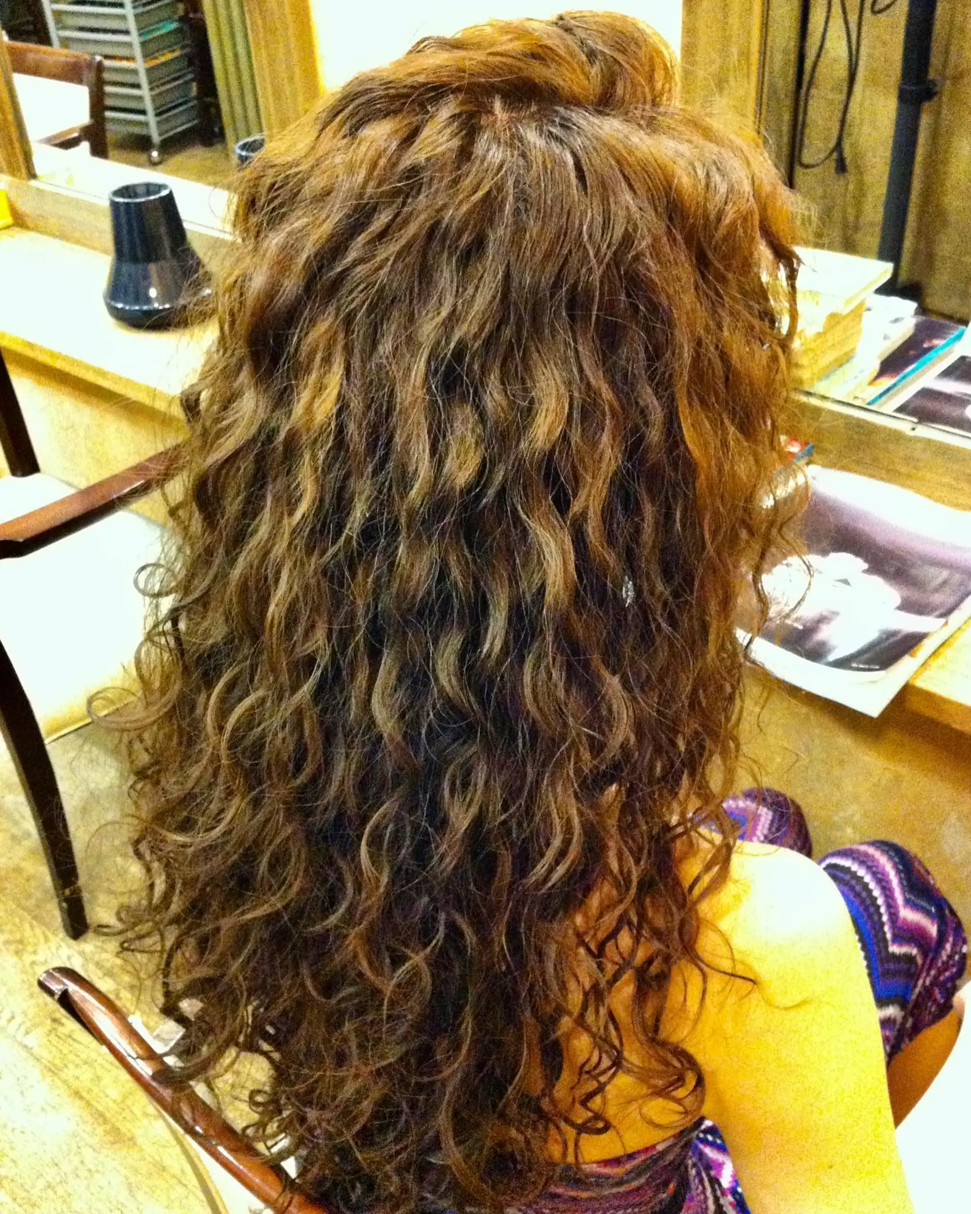 Long hair styled into full, defined spiral perm curls with natural texture and warm golden brown dimension. 1