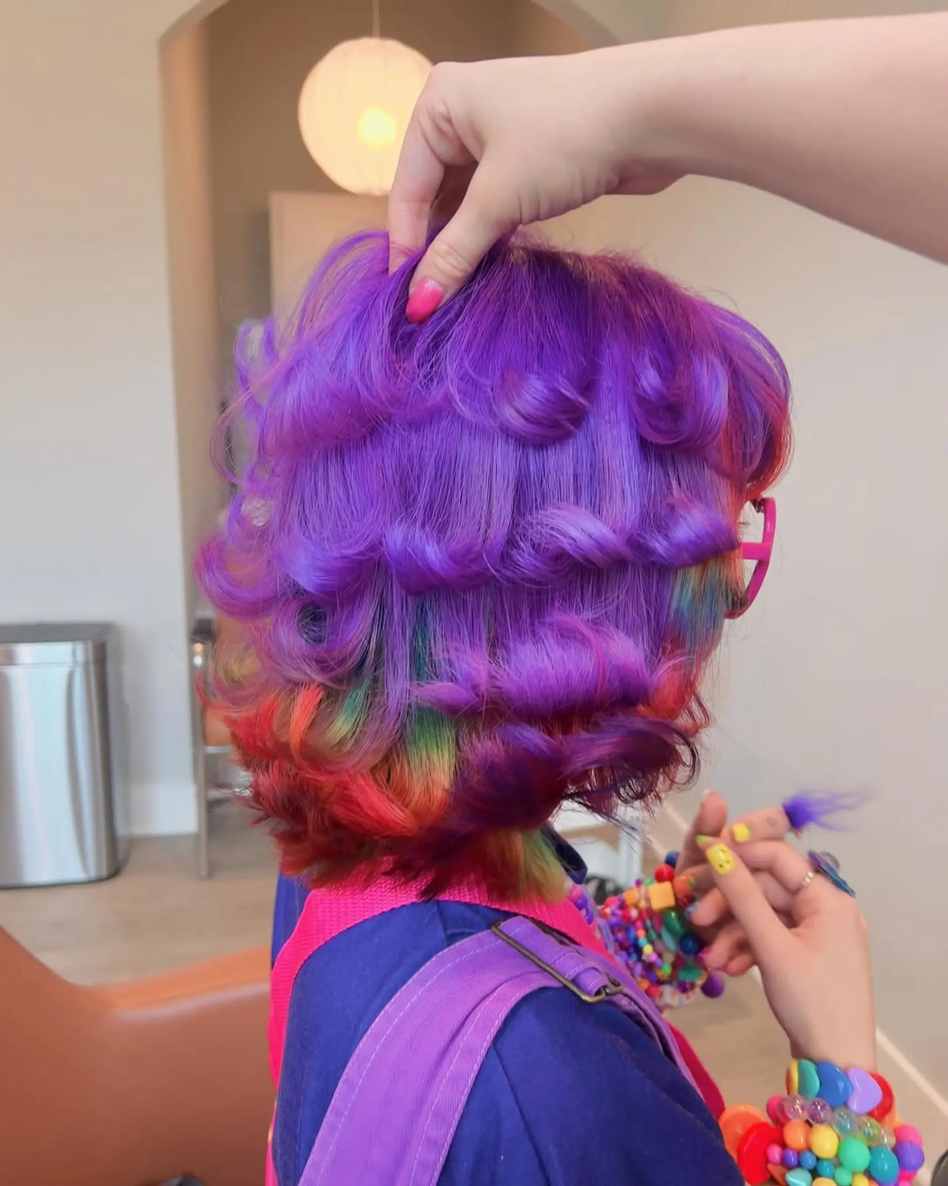 A fun, short curly bob style with bright purple hair on top and a surprise rainbow underlayer, creating a peekaboo effect when the hair moves, offering a dynamic and playful look. 1