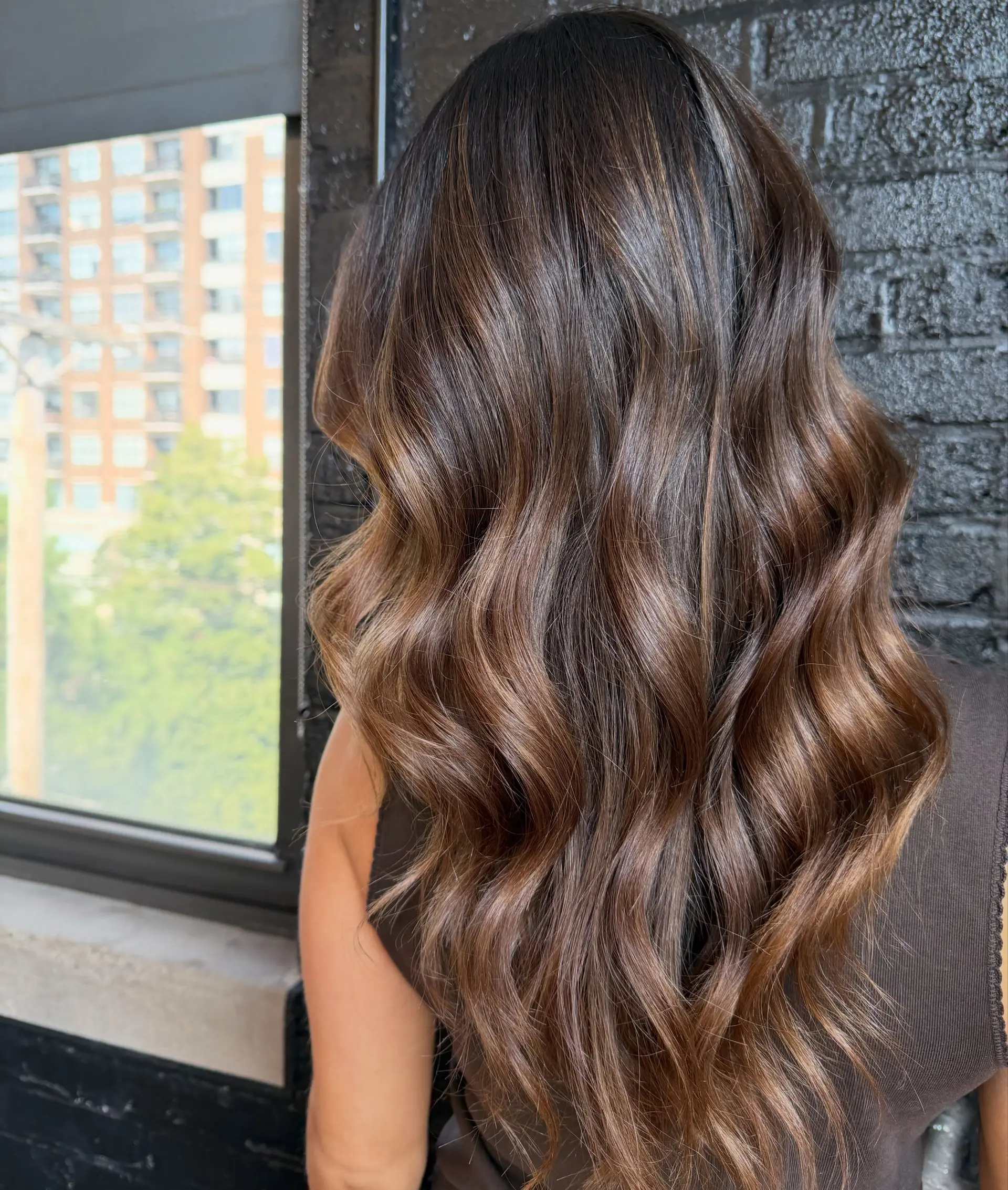 Long dimensional brunette hair styled in glossy, soft waves with a seamless root melt into warm caramel and light brown tones. 1