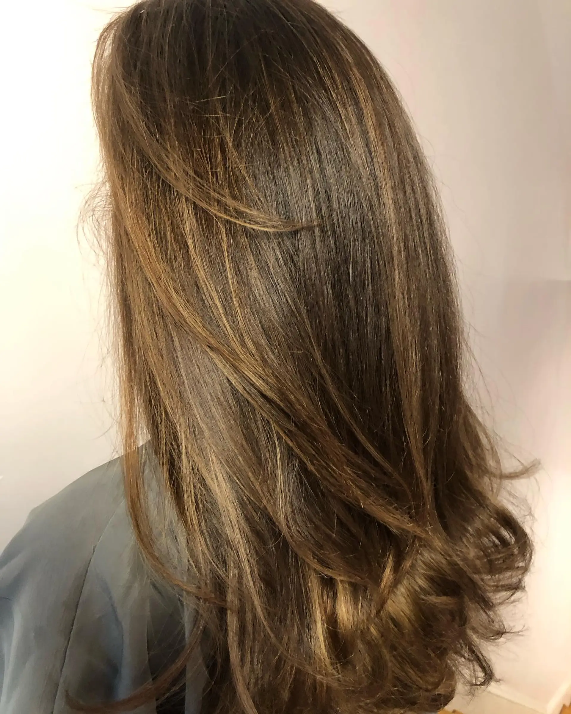 Long brunette hair cut with soft, cascading layers and painted with subtle caramel balayage, finished with a smooth blowout and curled ends. 1
