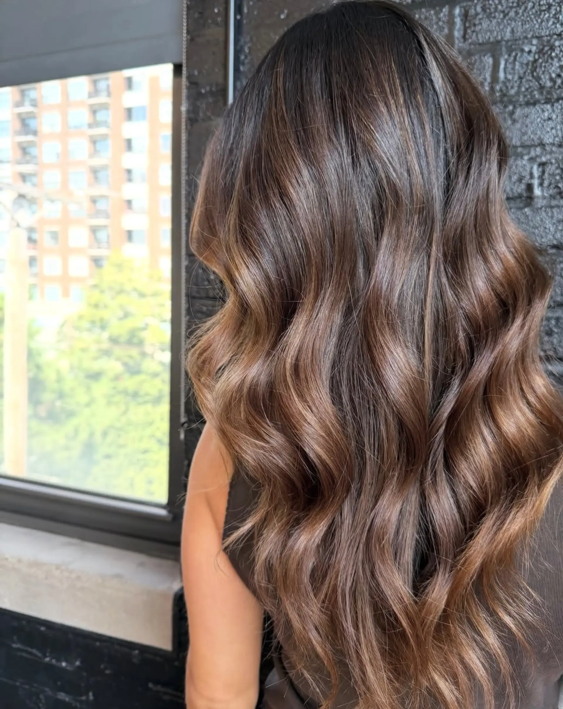 Long dimensional brunette hair styled in loose, glossy waves featuring soft caramel balayage highlights. 1
