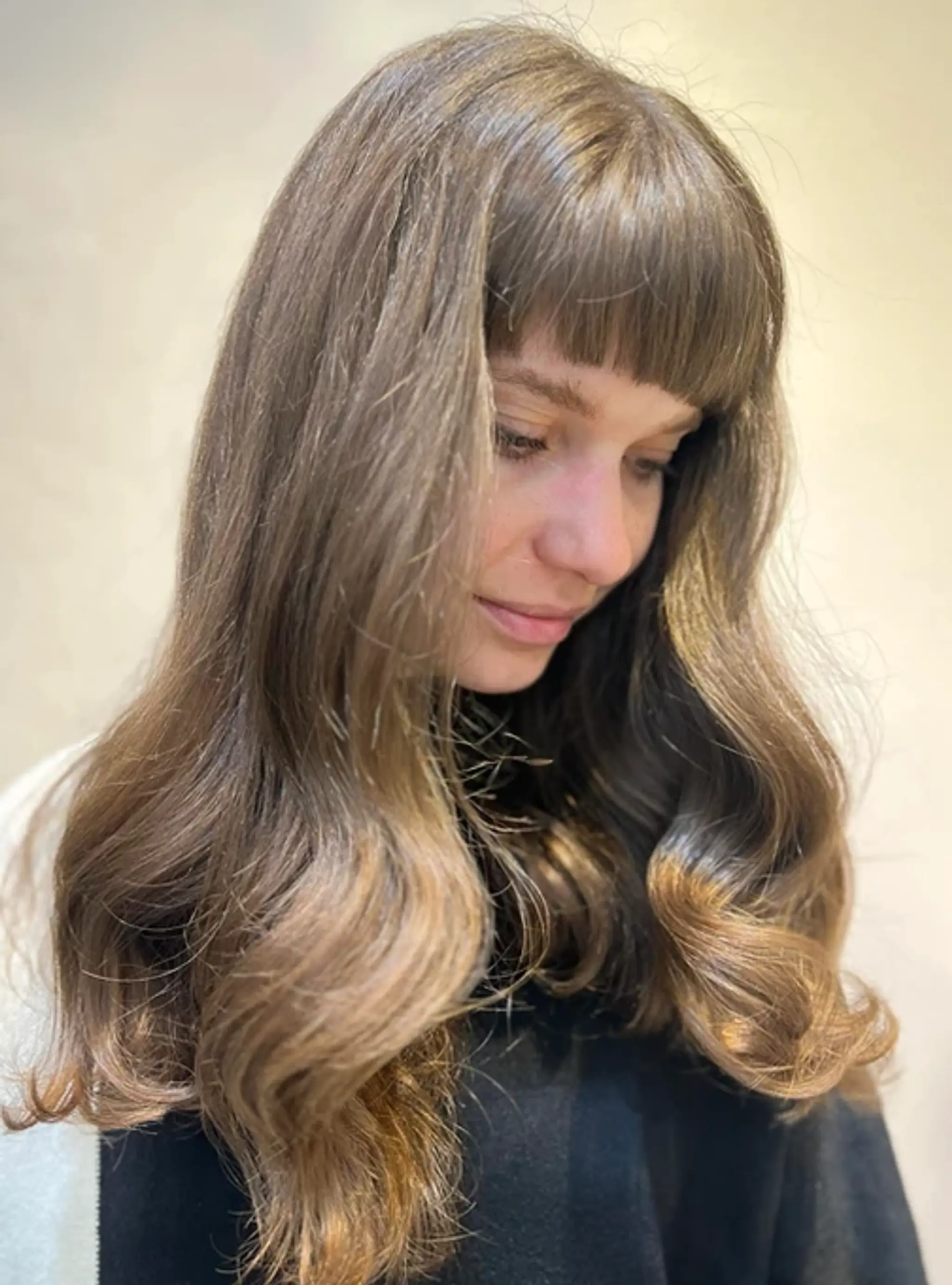 Long, wavy light brown hair with golden highlights, featuring a thick blunt fringe and naturally layered waves.