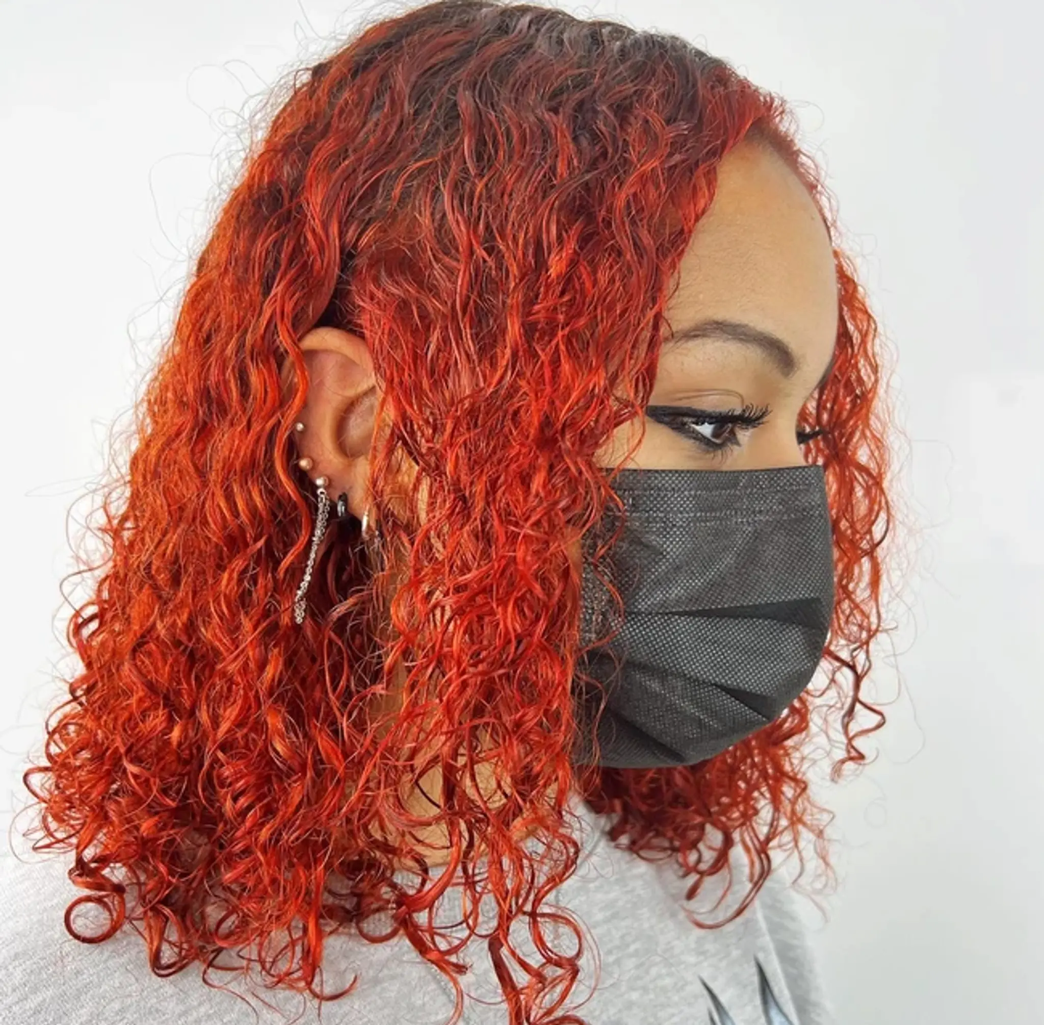 Medium-length, curly hair featuring a vibrant fiery red color with a subtle side part and tight, natural curls that fall freely, highlighting the rich red hue.