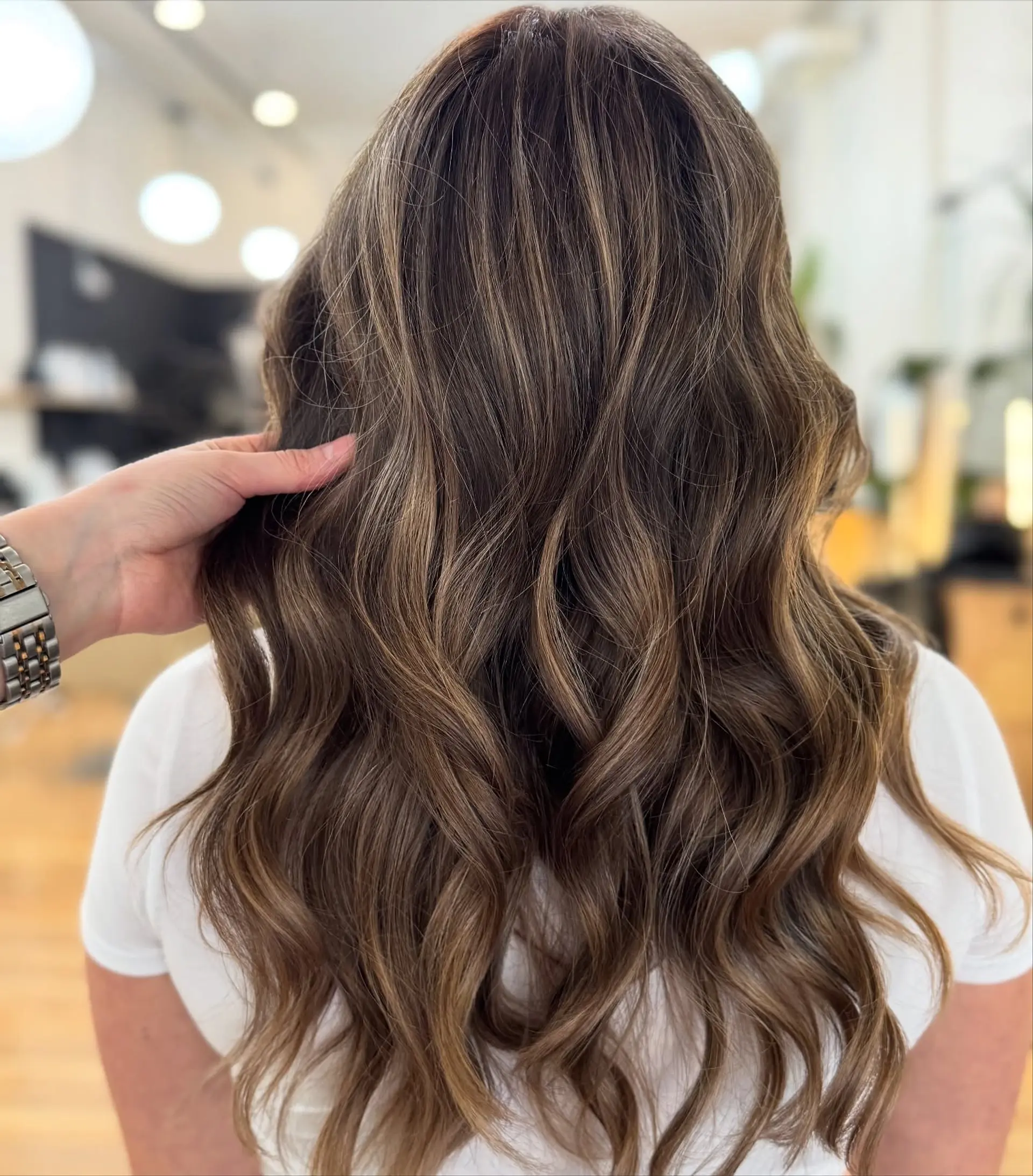 Long dimensional brunette hair styled in soft, flowing textured waves with seamless warm highlights. 1