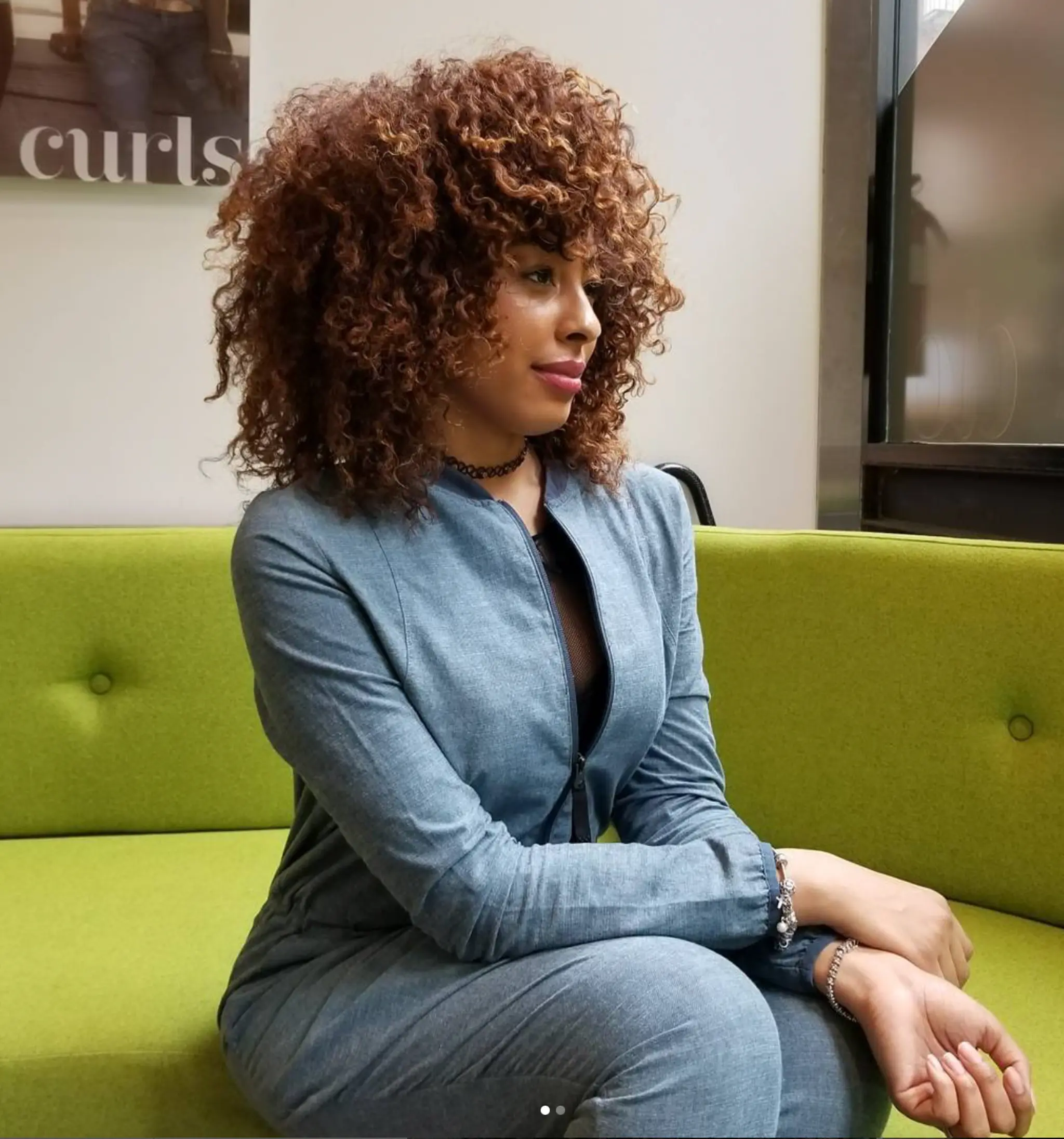 This hairstyle features voluminous, natural curls cascading around the head for a lively look. The curls are medium-length and have a uniform, defined shape, creating a bouncy, textured effect. The hair color is a rich auburn, enhancing the vibrancy and dimension of the curls. Styled with a subtle side part, the curls frame the face beautifully.