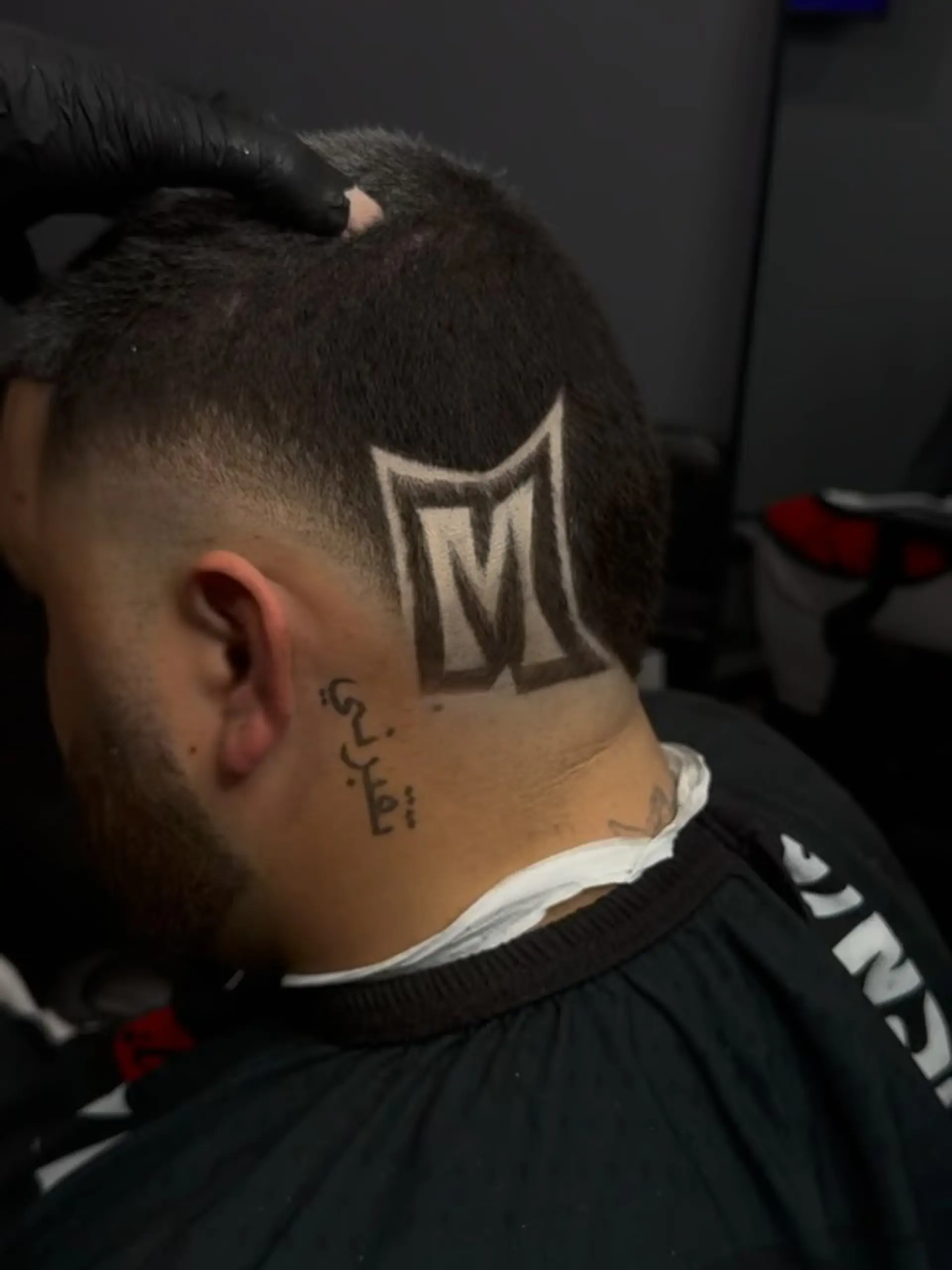 A short fade haircut featuring an intricate, sharp 3D letter 'M' shaved into the side of the head. 1
