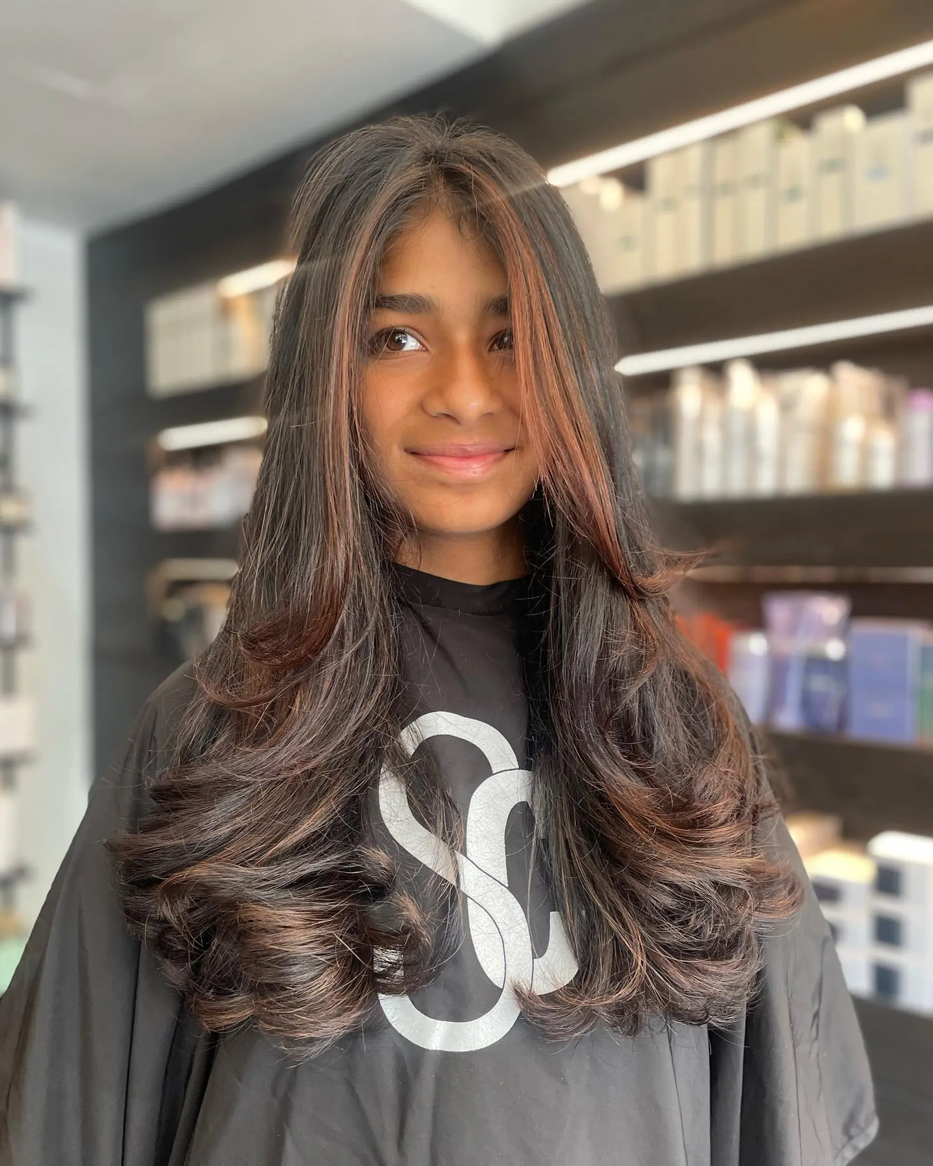 Long layered dark hair styled into a bouncy blowout with warm face-framing highlight accents. 1