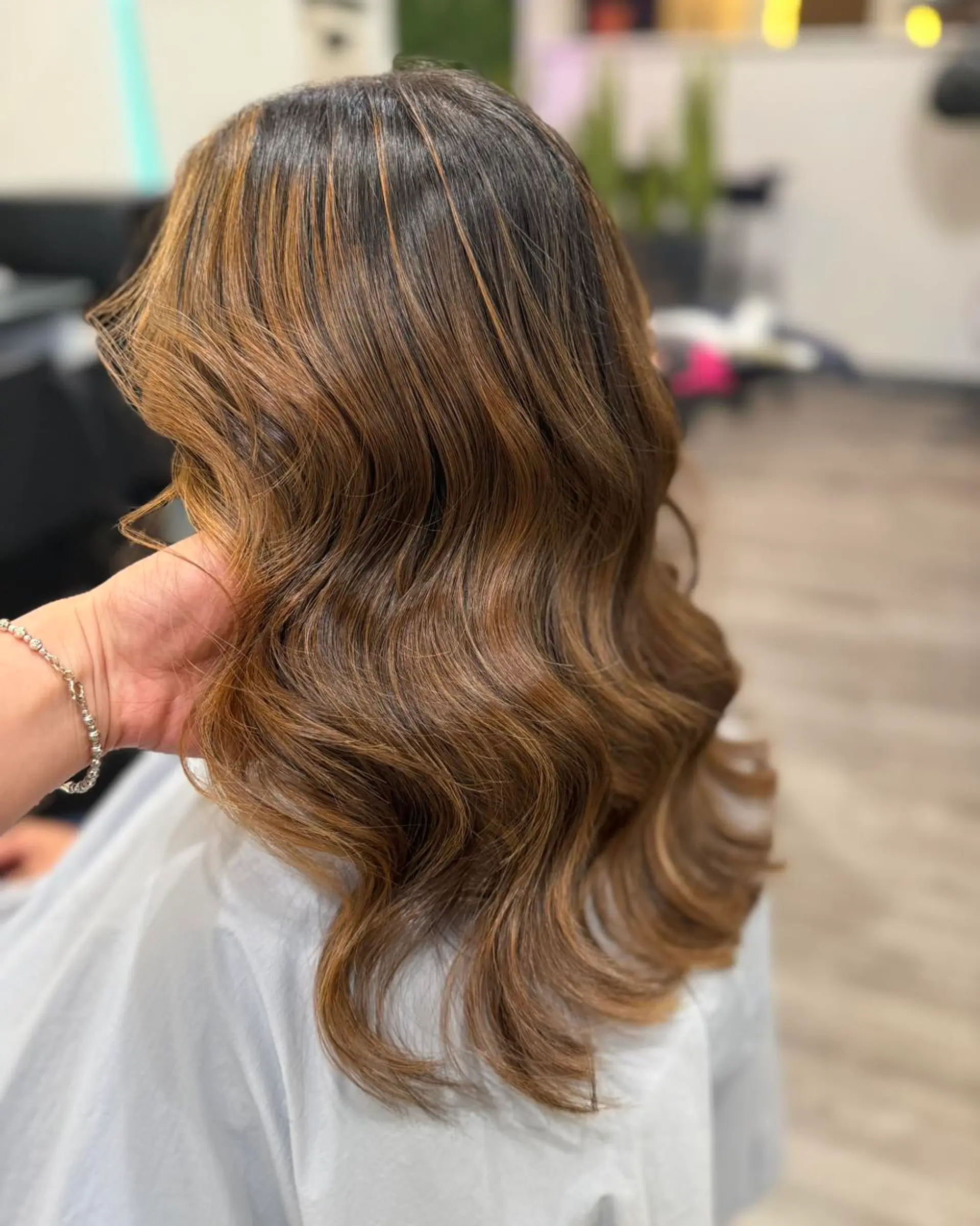 Medium-length dimensional brunette hair styled in soft, polished waves with warm caramel highlights. 1